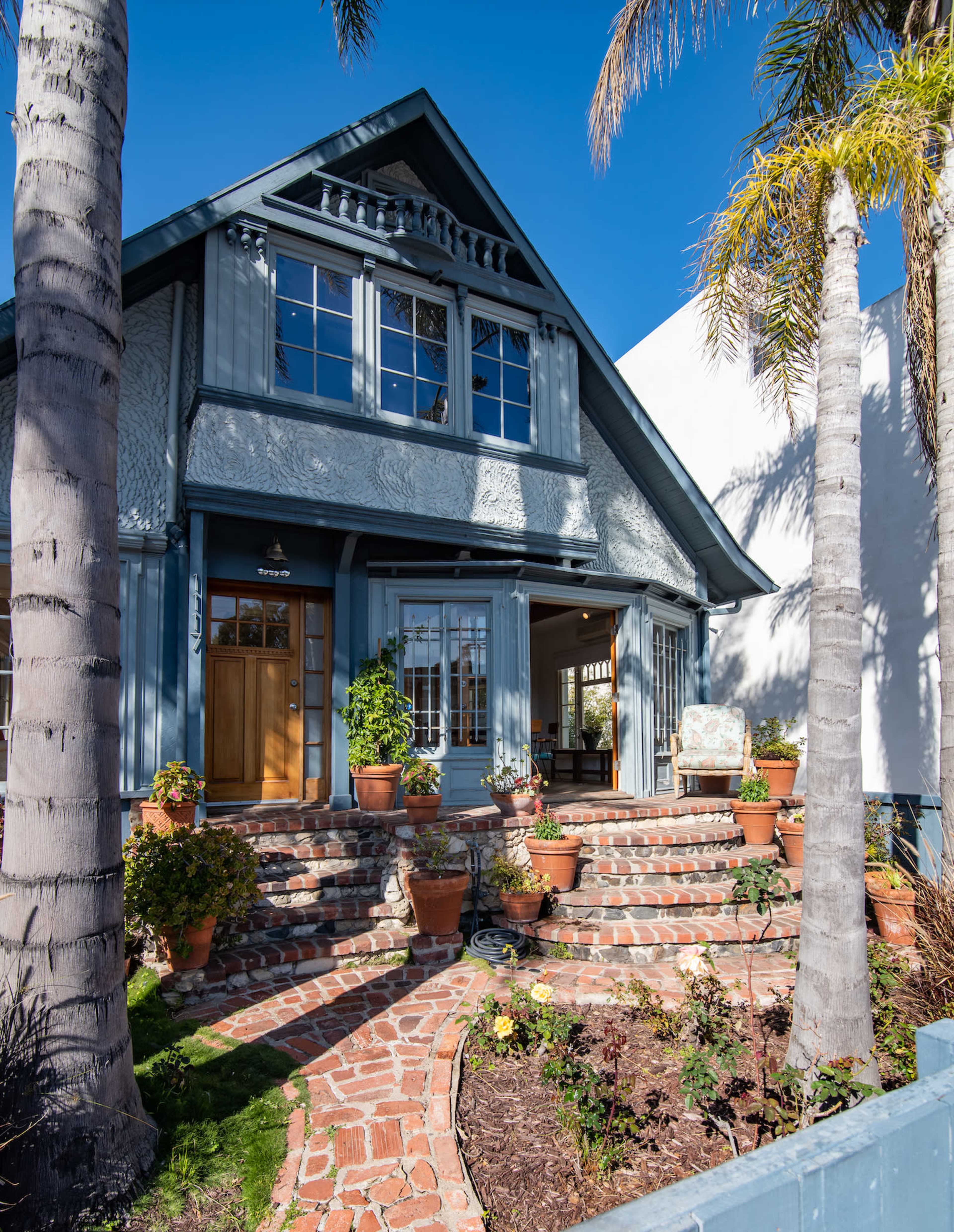 A blue bungalow with a landscaped front yard is framed by palm trees and features a brick pathway leading to the entrance.
