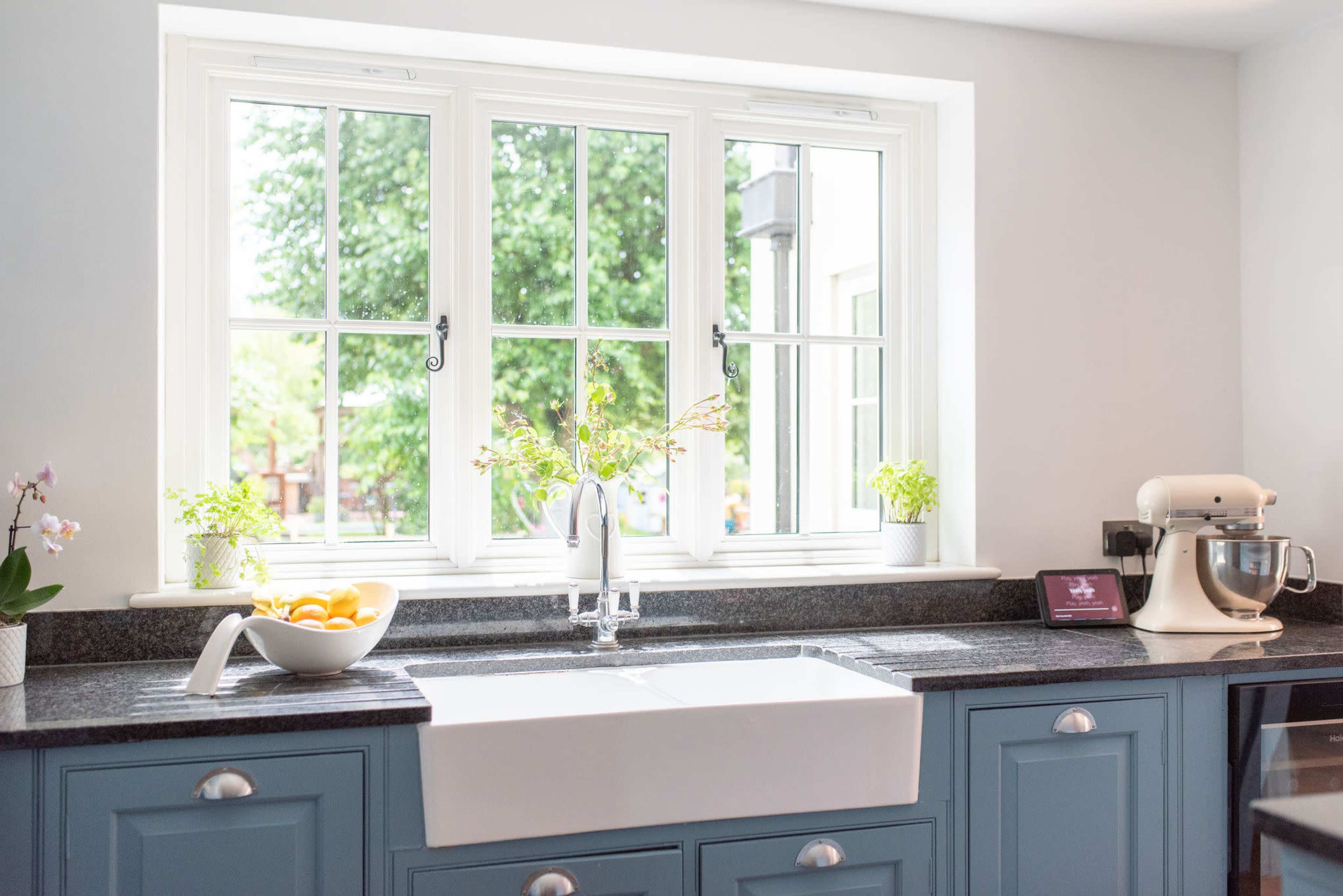 A clean kitchen features a large farmhouse sink, granite countertops, and a window surrounded by potted plants, with a bowl of lemons and a kitchen mixer nearby.