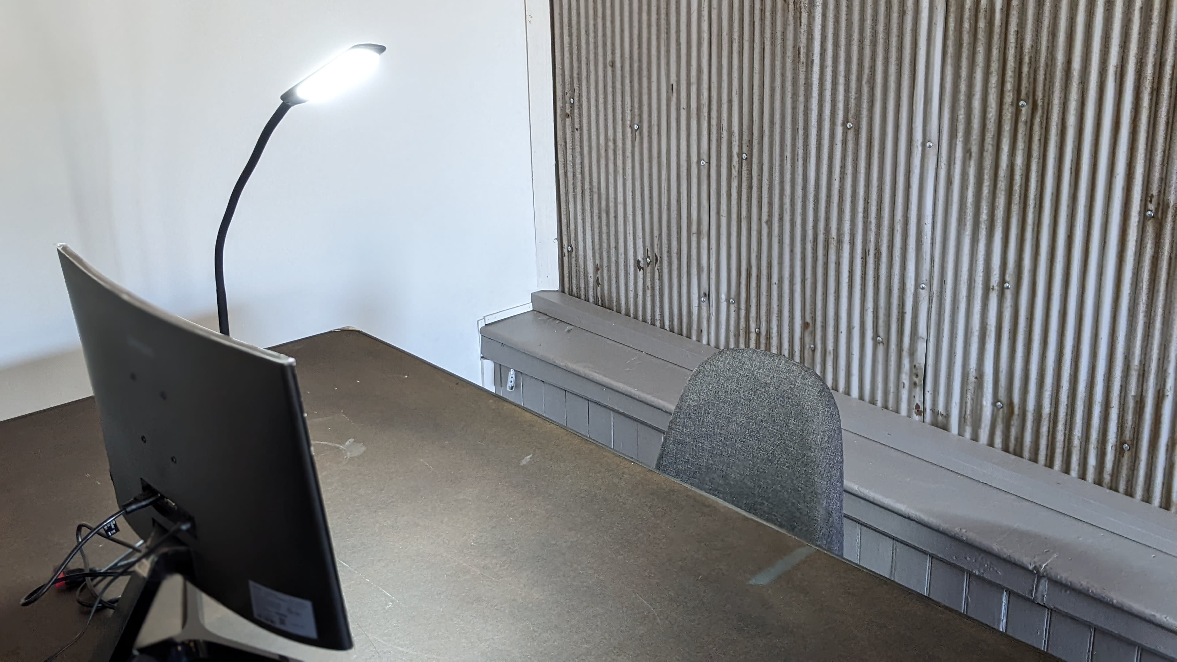 A desk with a computer monitor and a chair is positioned near a corrugated metal wall, with a lamp providing light.