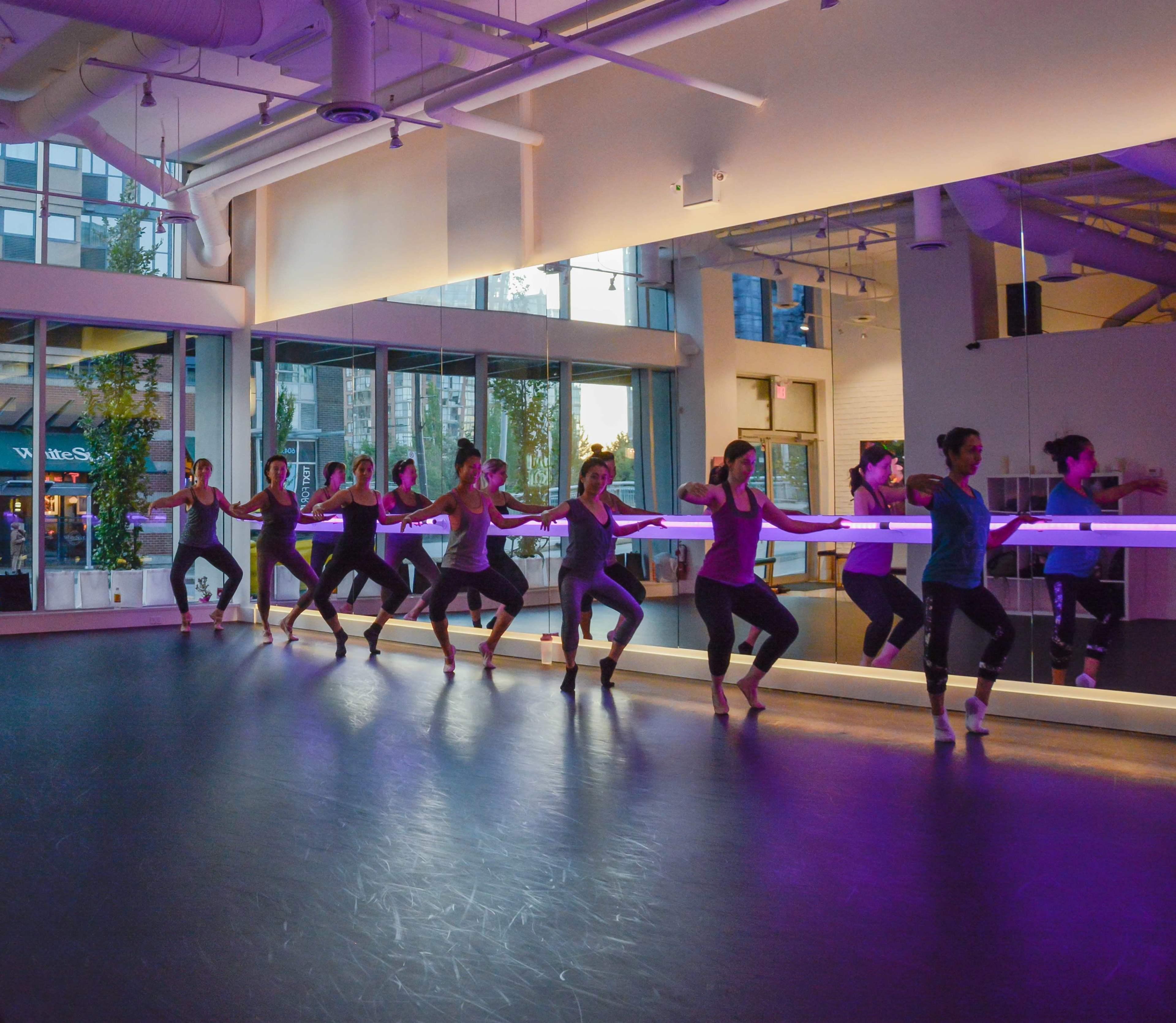 A group of dancers practices in a brightly lit studio with large windows and purple lighting.