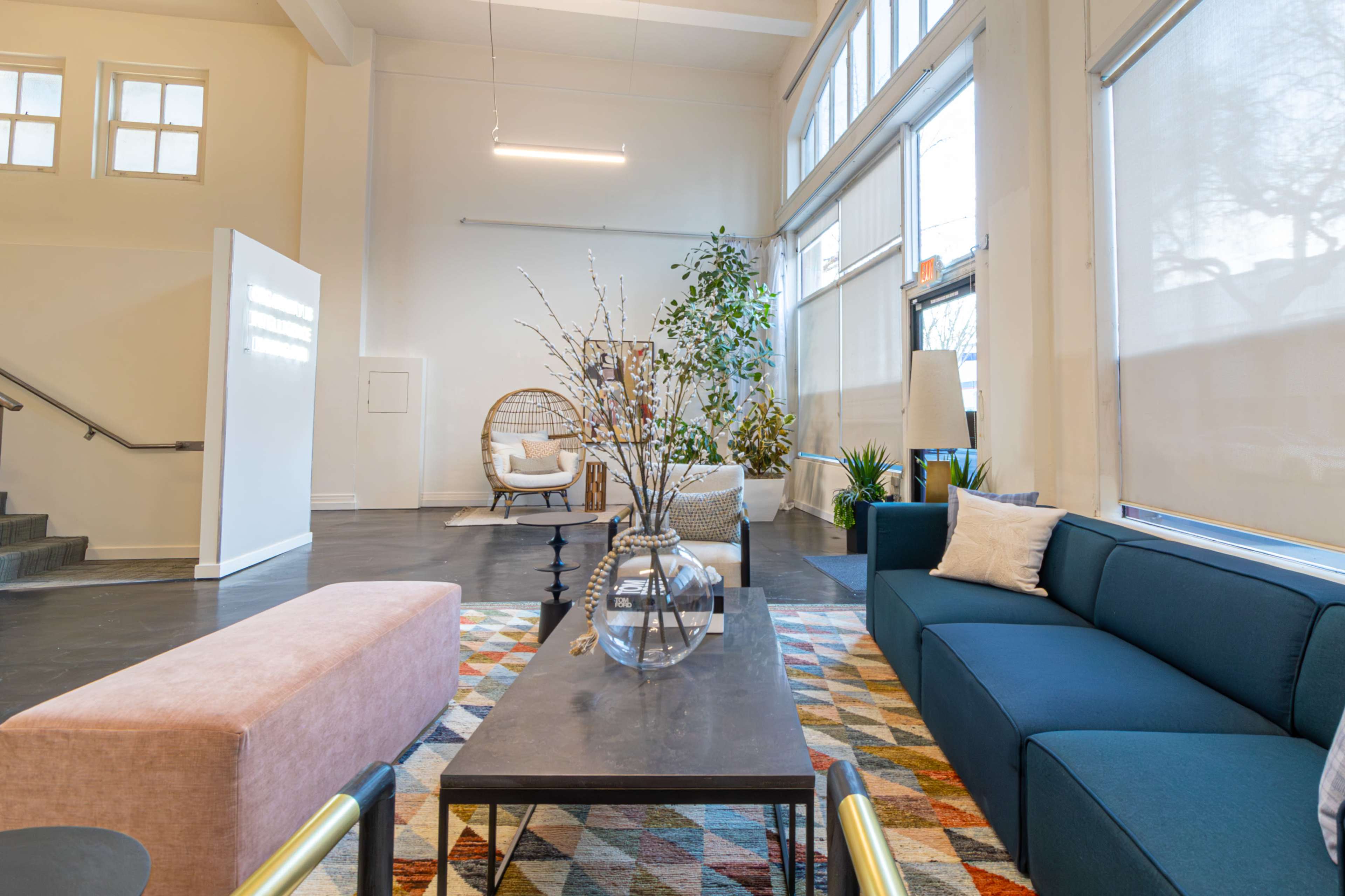 The image shows a modern living space furnished with a blue sofa, a pink bench, a coffee table with a vase, and various plants, illuminated by natural light from large windows.