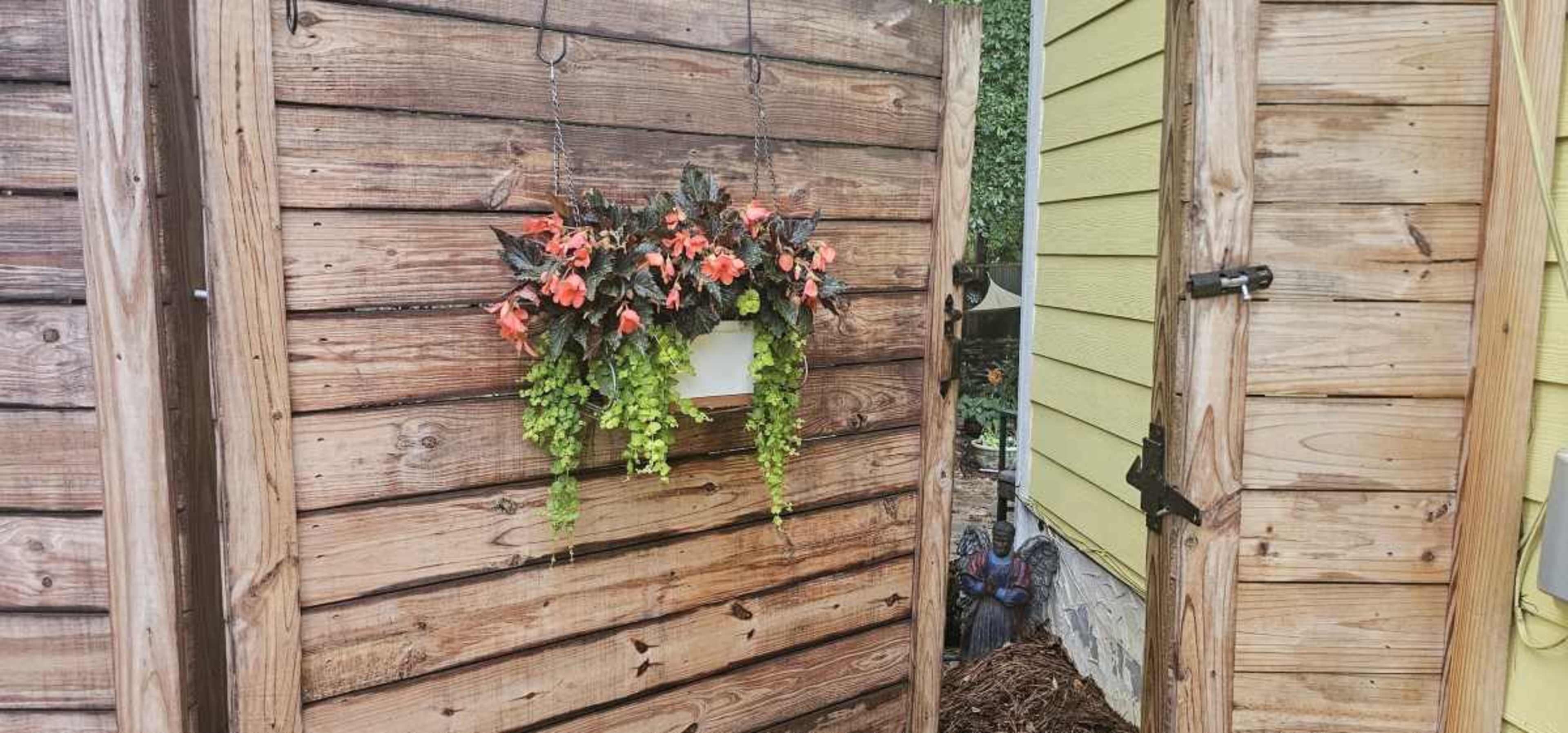 A wooden gate opens to reveal a planter with vibrant flowers hanging on a wall beside a house.
