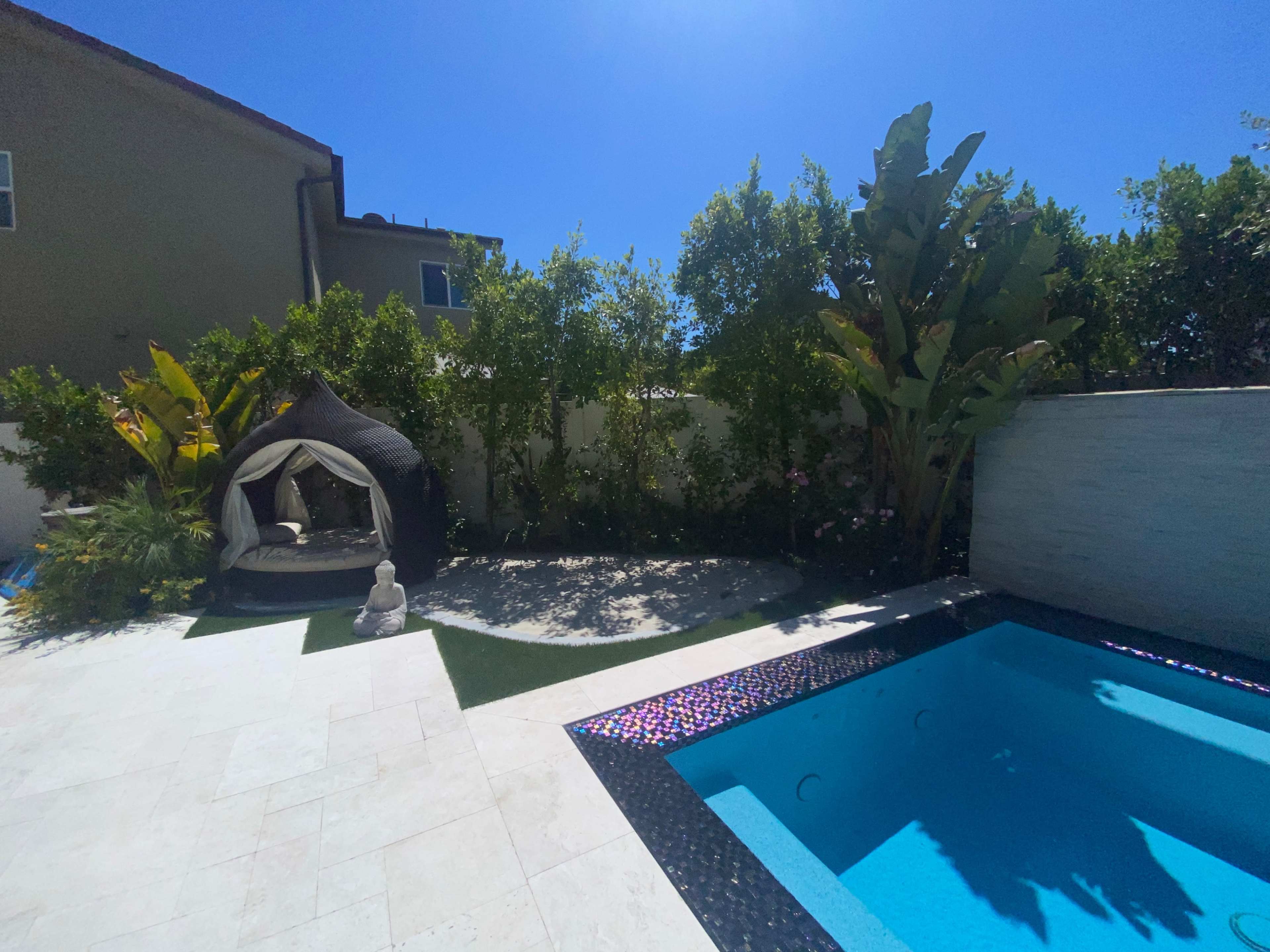 A backyard with a swimming pool, a hut structure surrounded by lush greenery, and a decorative statue nearby.