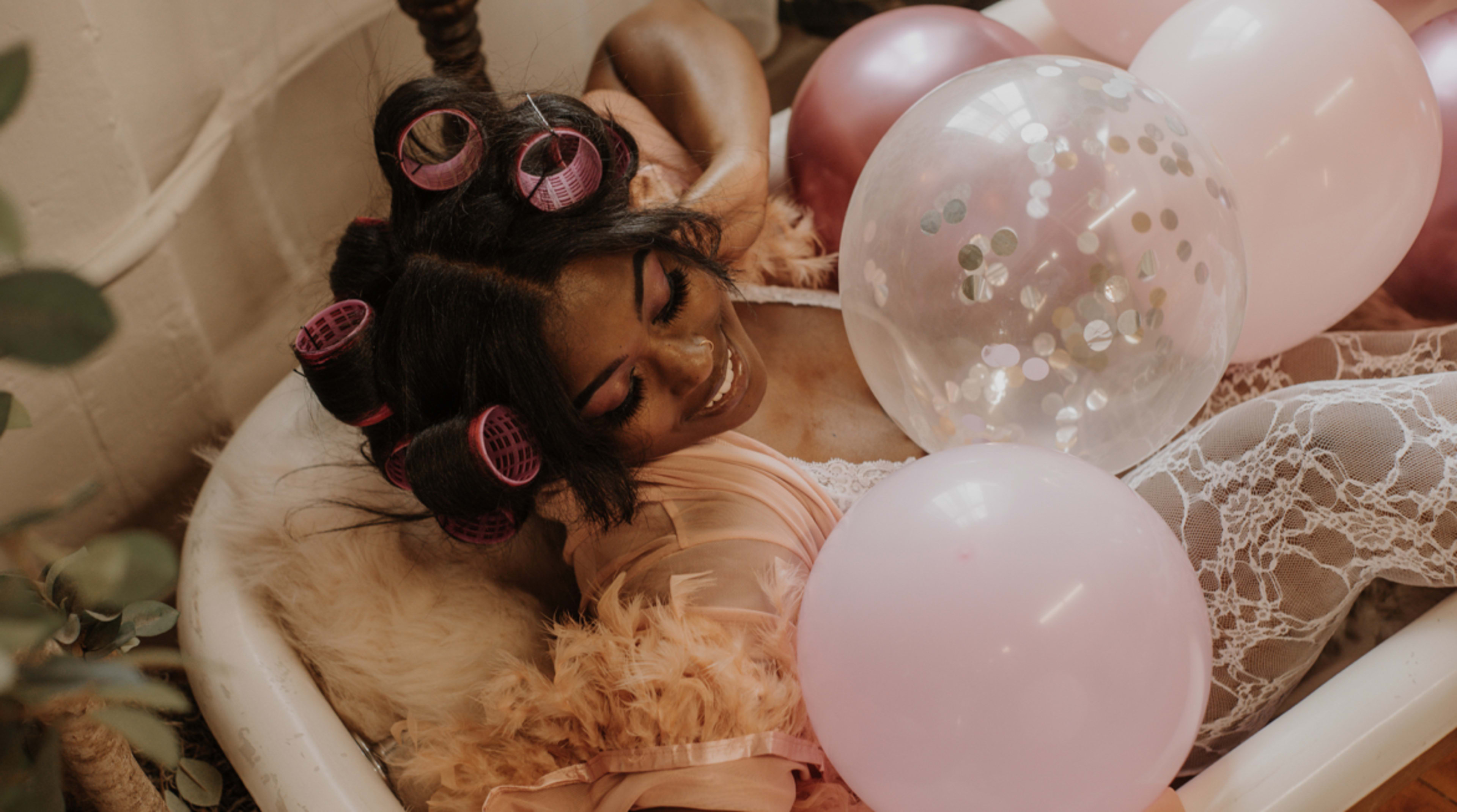 A boudoir photoshoot featuring a woman laying in a bathtub surrounded by rustic beige and pink balloons.