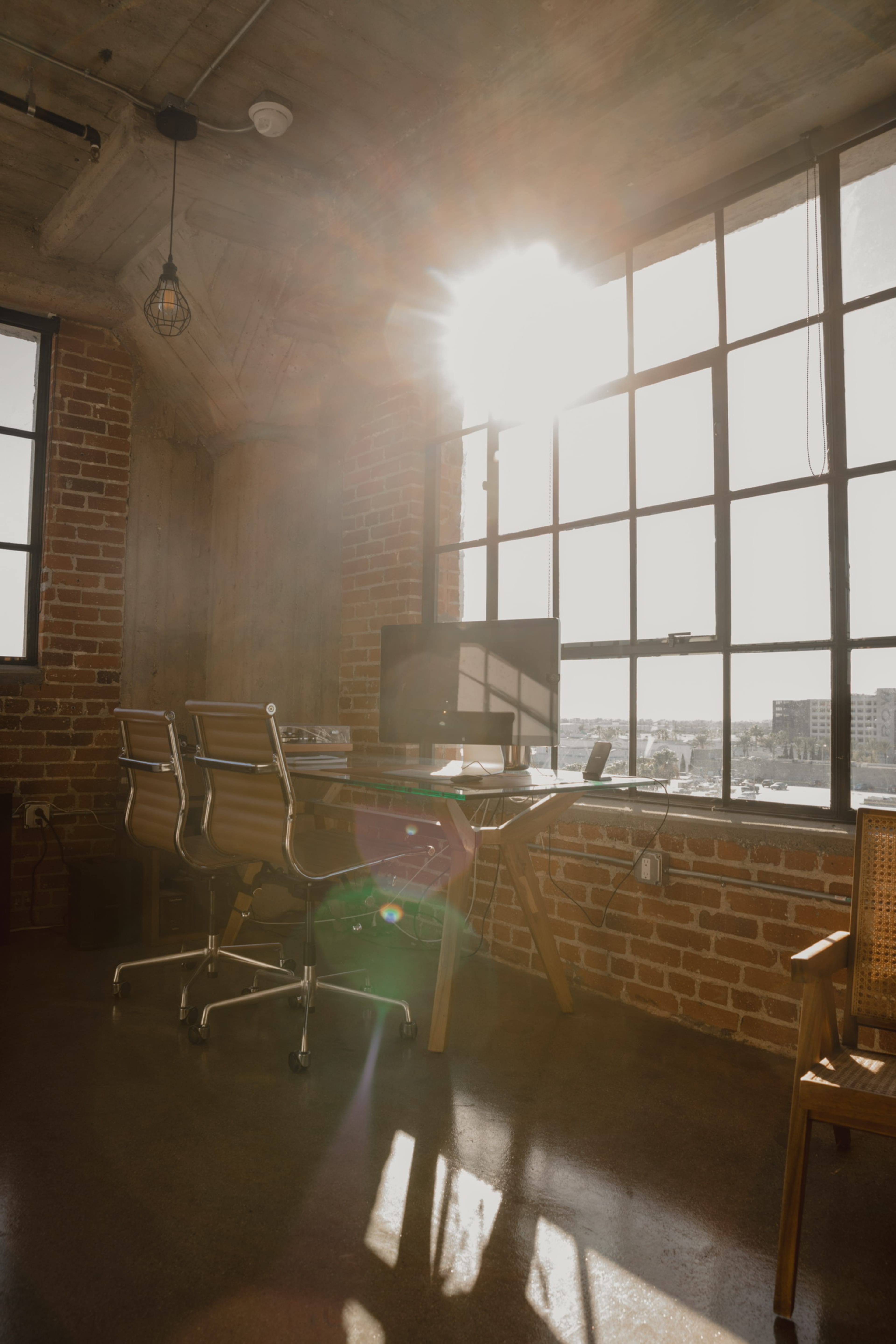A sunlit office features a wooden desk with two chrome chairs, large windows with brick walls, and a computer setup.
