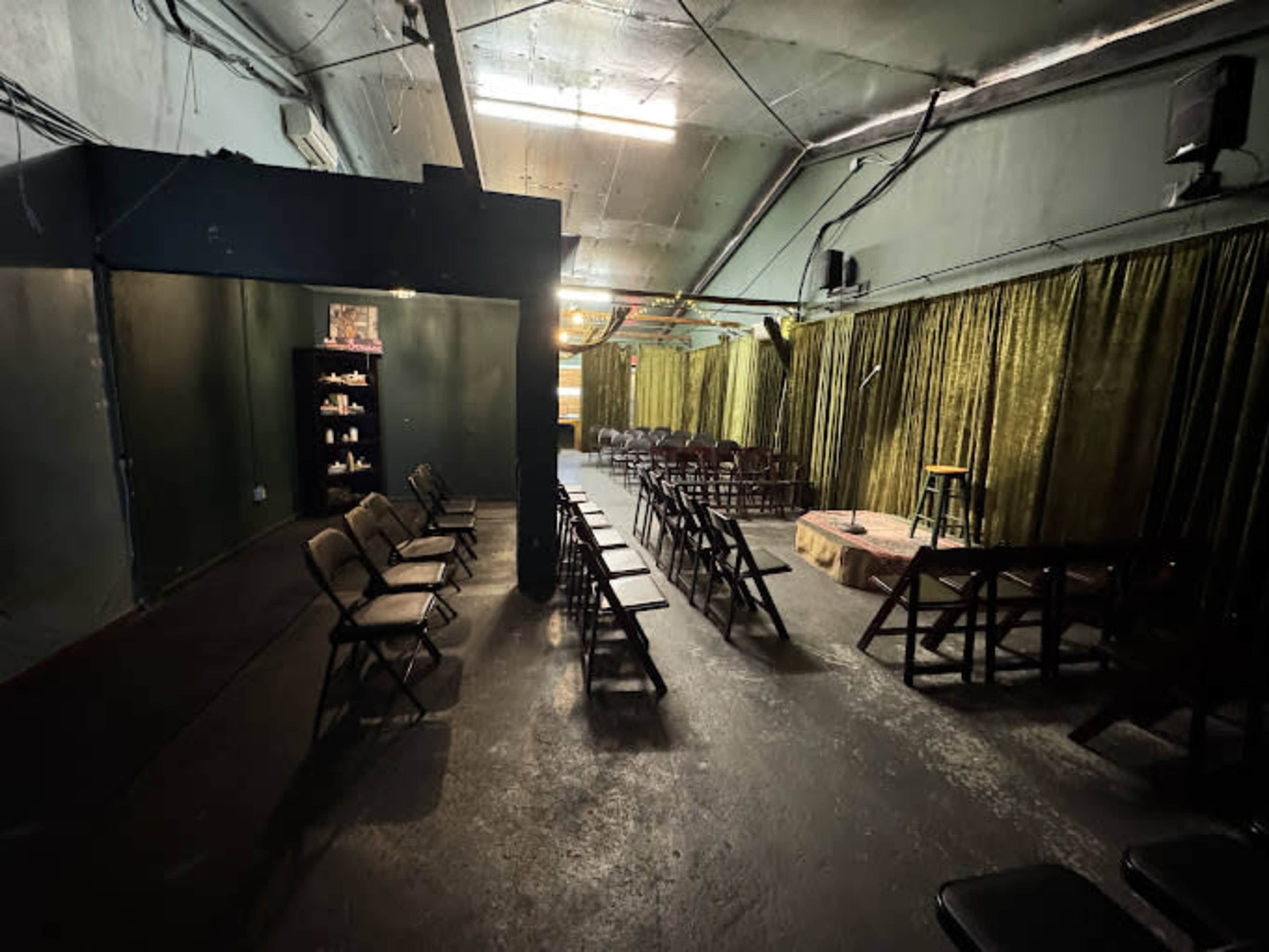 The image shows an empty performance space with rows of folding chairs facing a small raised stage, surrounded by green curtains.