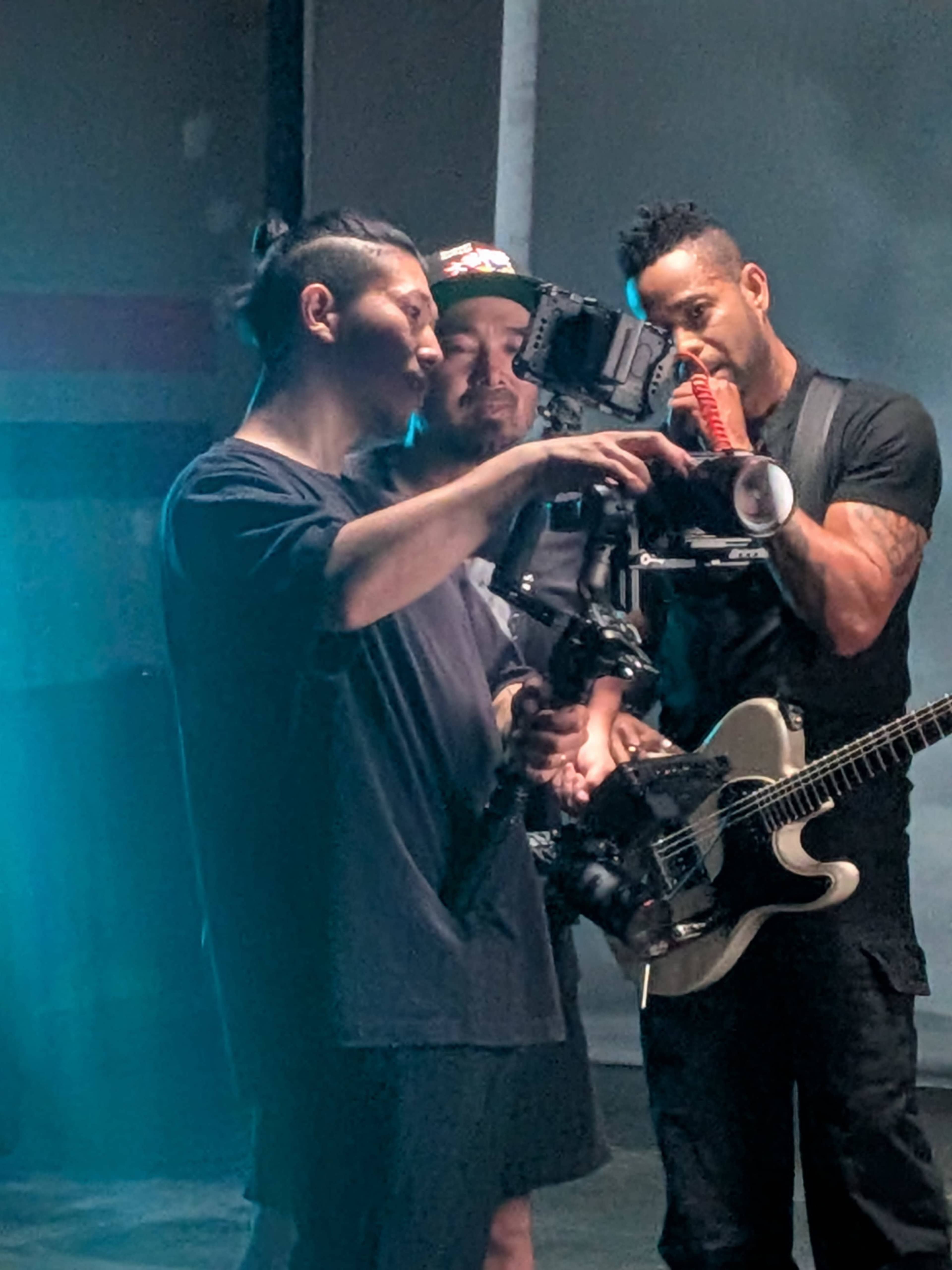 Three individuals are engaged in a discussion while reviewing a camera setup, with one holding a guitar.