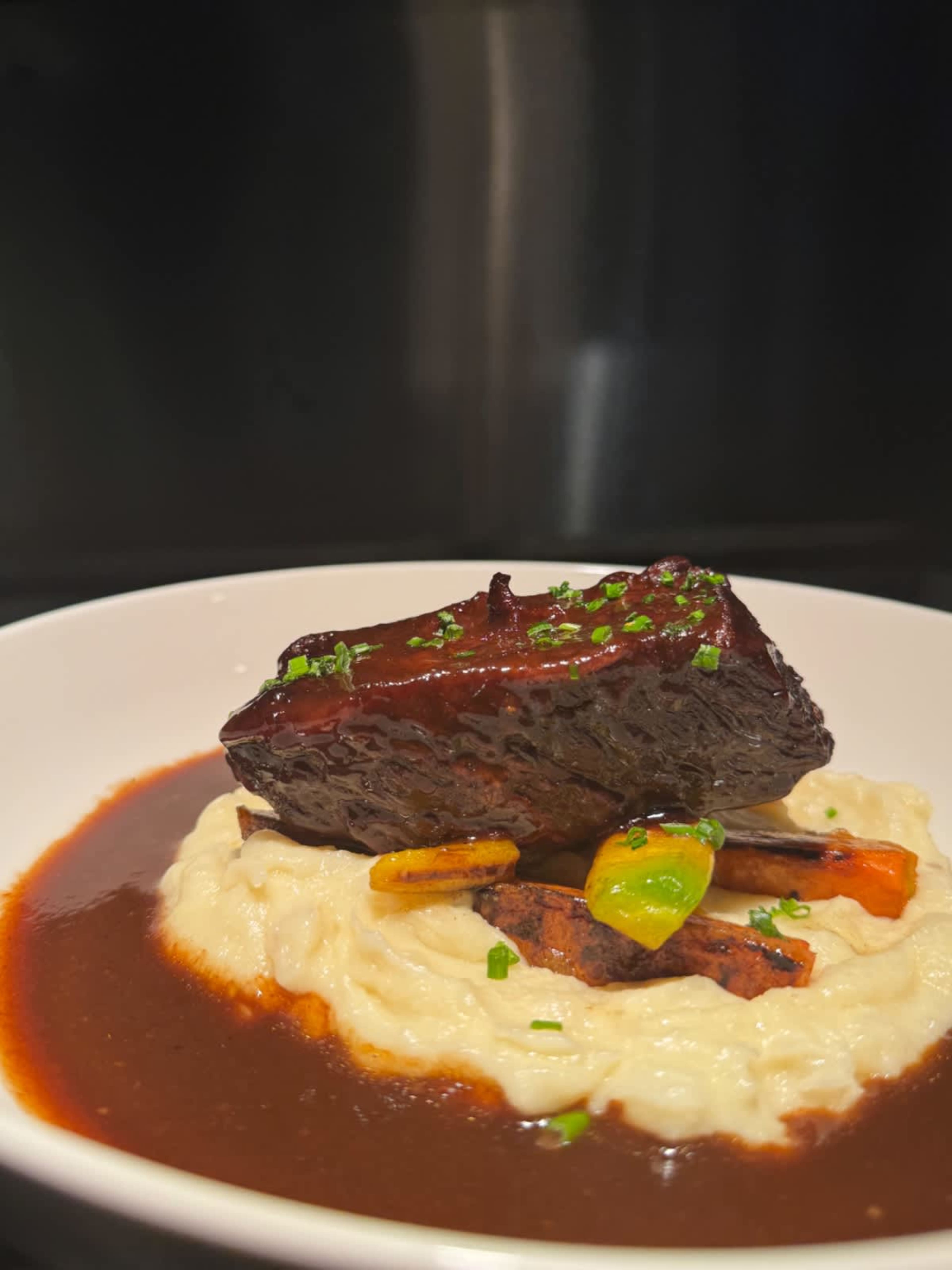 A piece of braised meat is topped with sauce and served over a bed of creamy mashed potatoes, garnished with green herbs and accompanied by colorful vegetables.