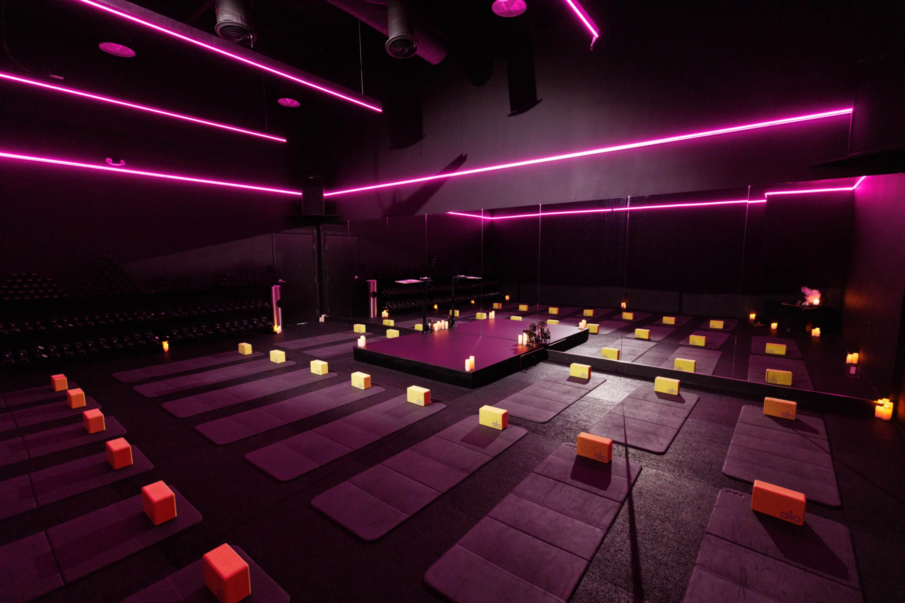 A dimly lit yoga studio features rows of exercise mats and blocks, illuminated by pink neon lights and candles.