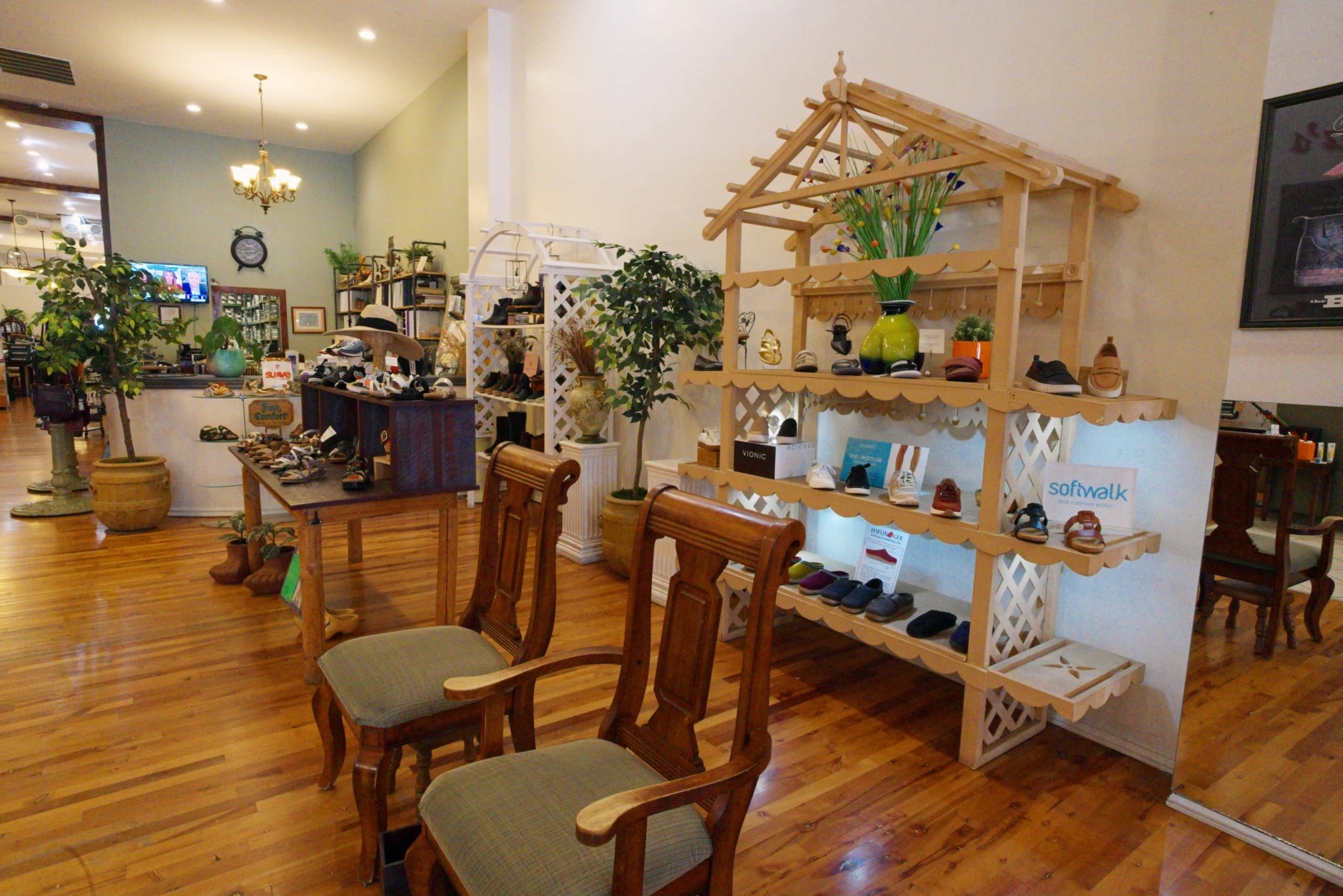 The image shows a retail shoe store interior featuring wooden shelves displaying various shoes, plants, and furniture, along with two wooden chairs in the foreground.