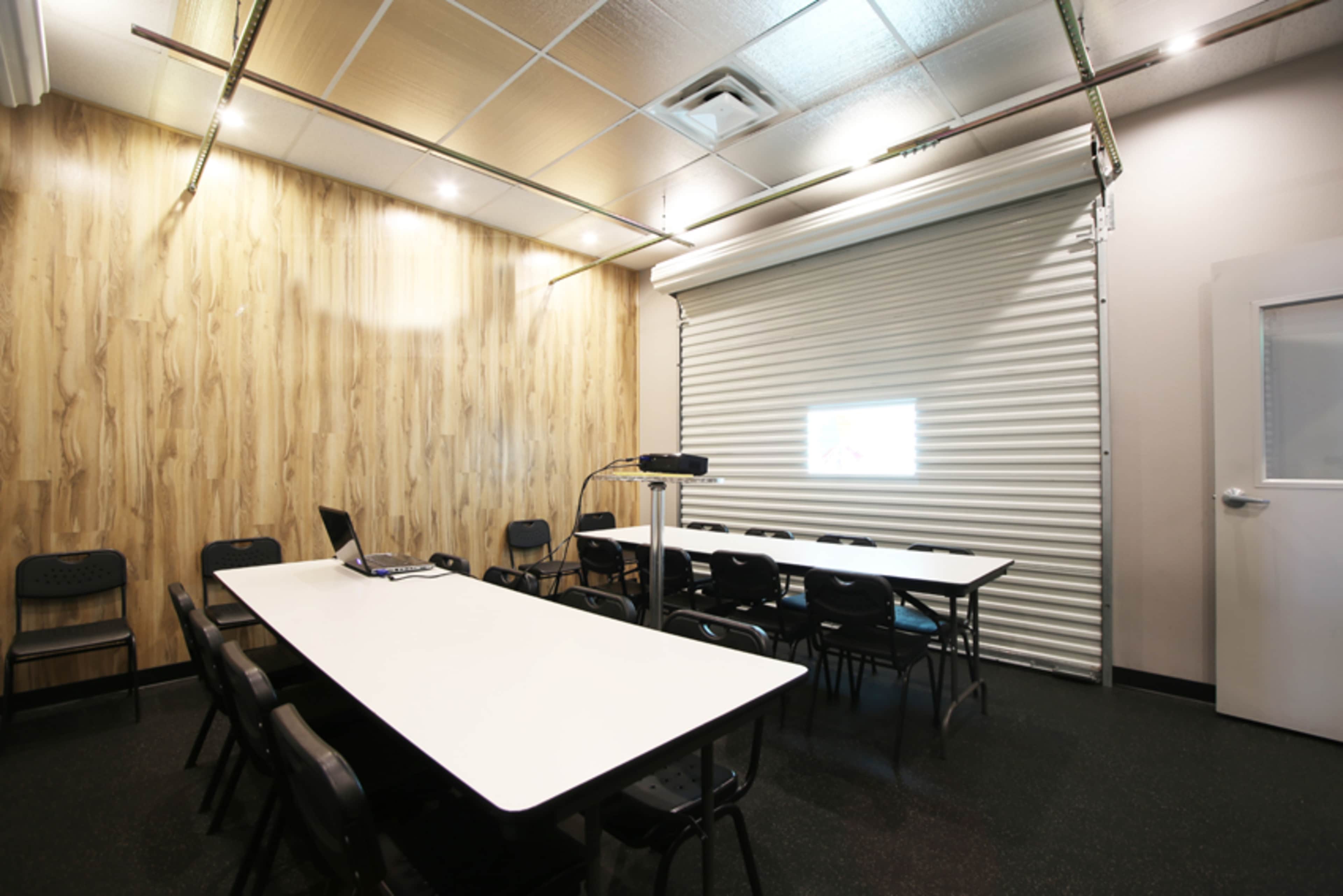 The image shows a meeting room with a long table, several black chairs, and a roll-up door at one end.