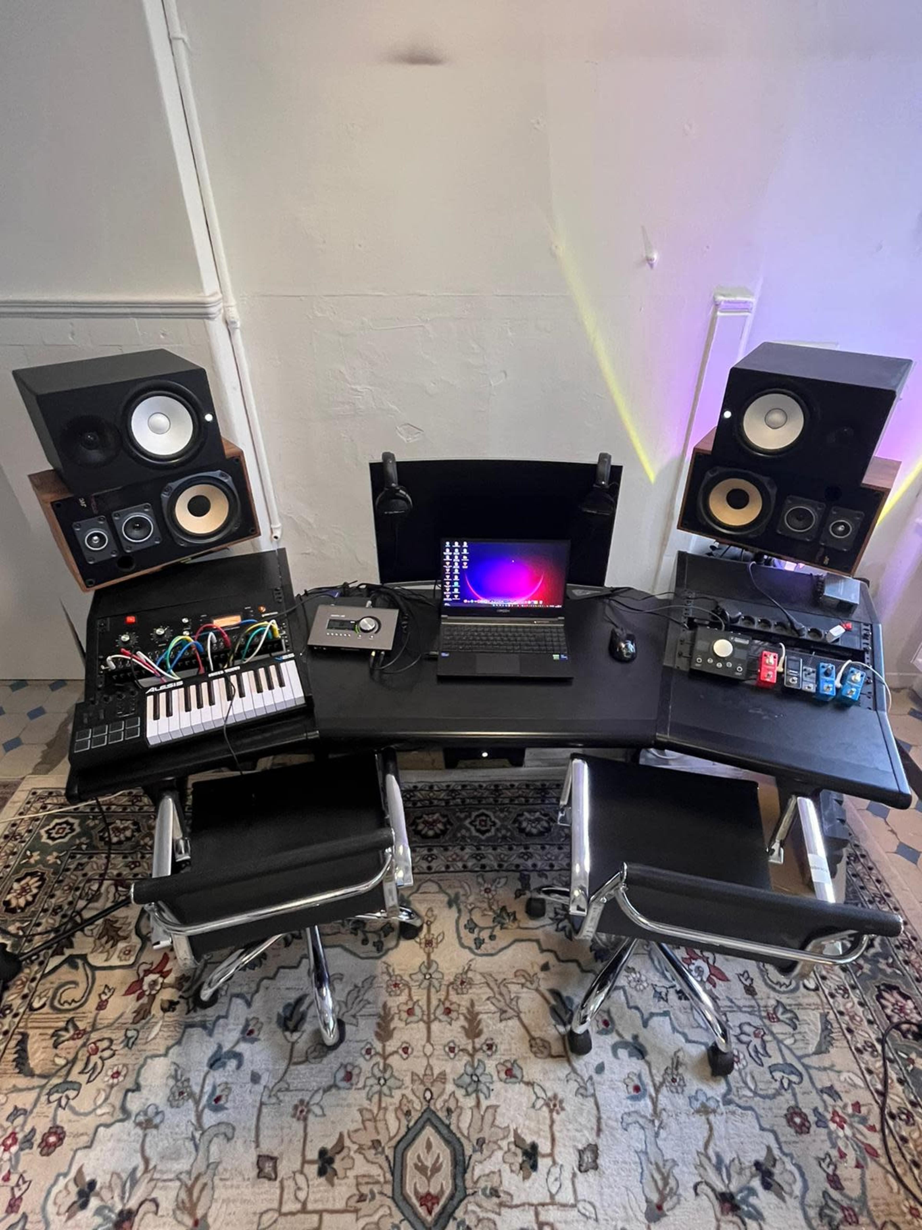 The image shows a music production setup featuring two studio monitor speakers, a laptop, a MIDI keyboard, and various audio equipment arranged on a desk with two chairs and a patterned rug.