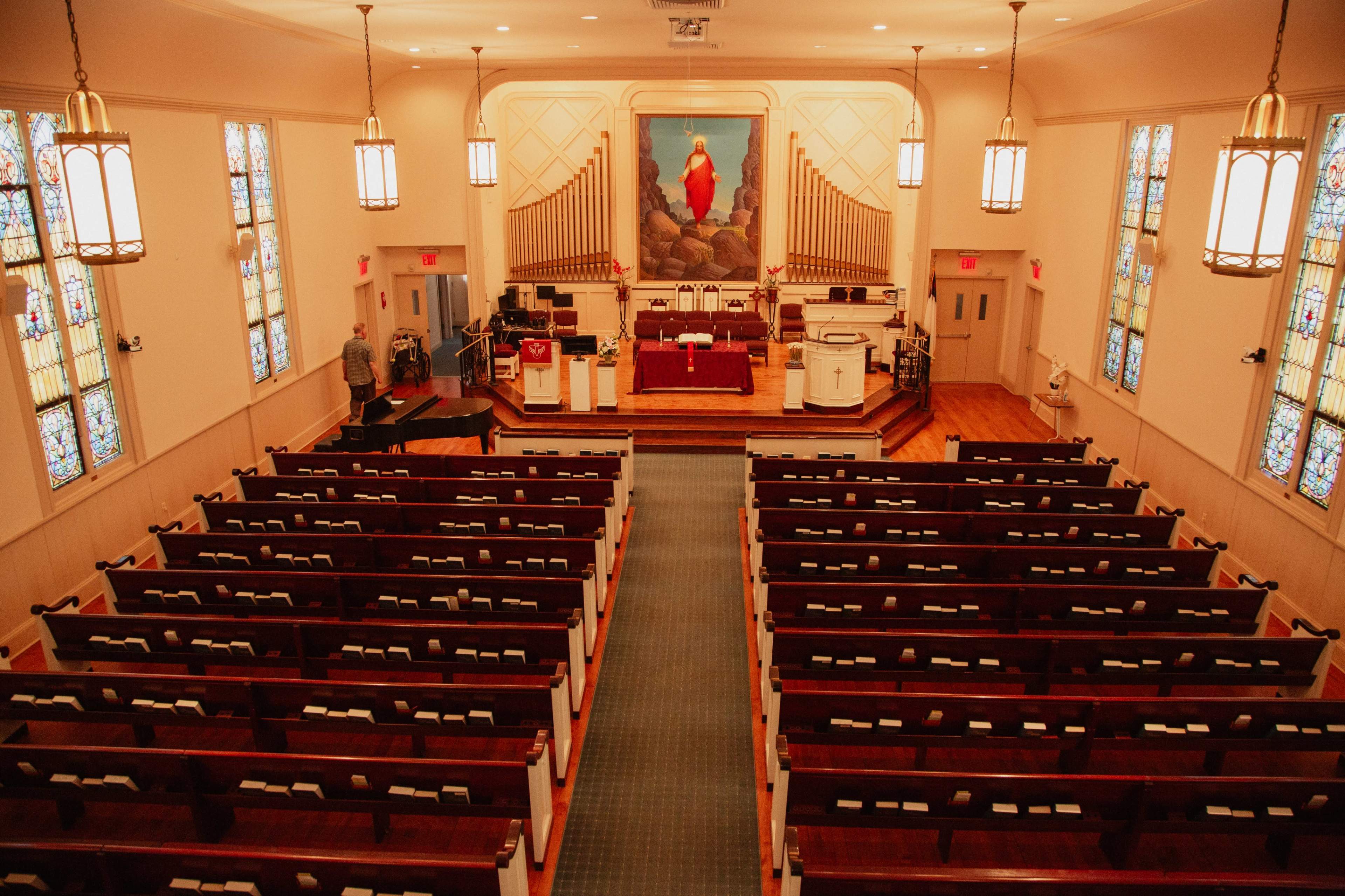 The image shows the interior of a church, featuring wooden pews arranged in rows facing an altar with a large painting and stained glass windows.