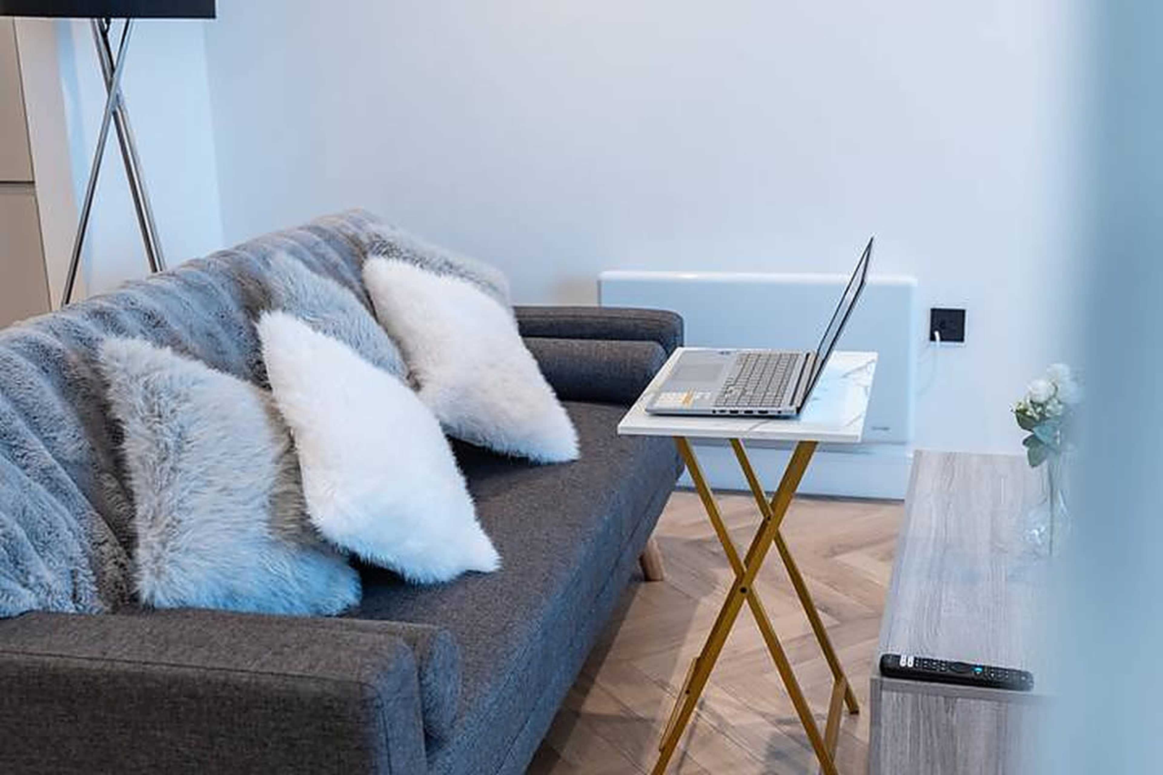 A gray sofa with fluffy white pillows is positioned next to a laptop on a small side table in a modern room.