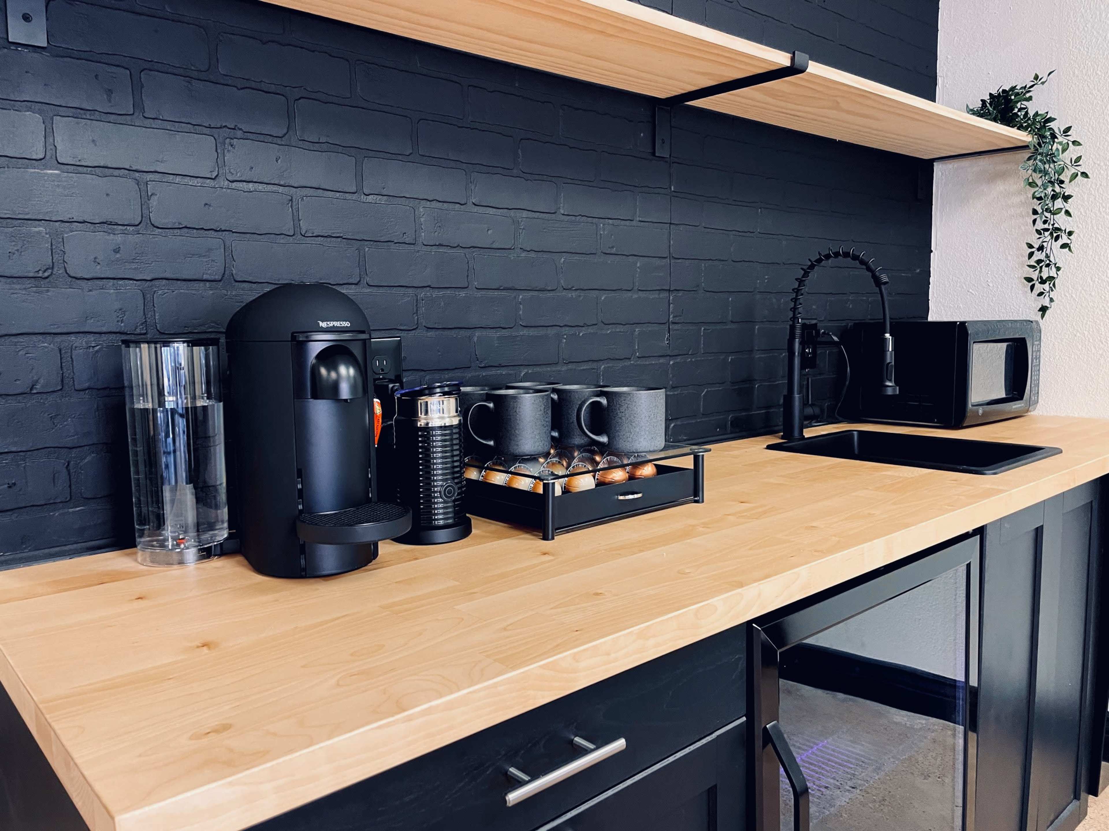 A modern kitchen countertop features a coffee machine, a coffee grinder, a shelf with mugs, and a microwave against a black brick wall.