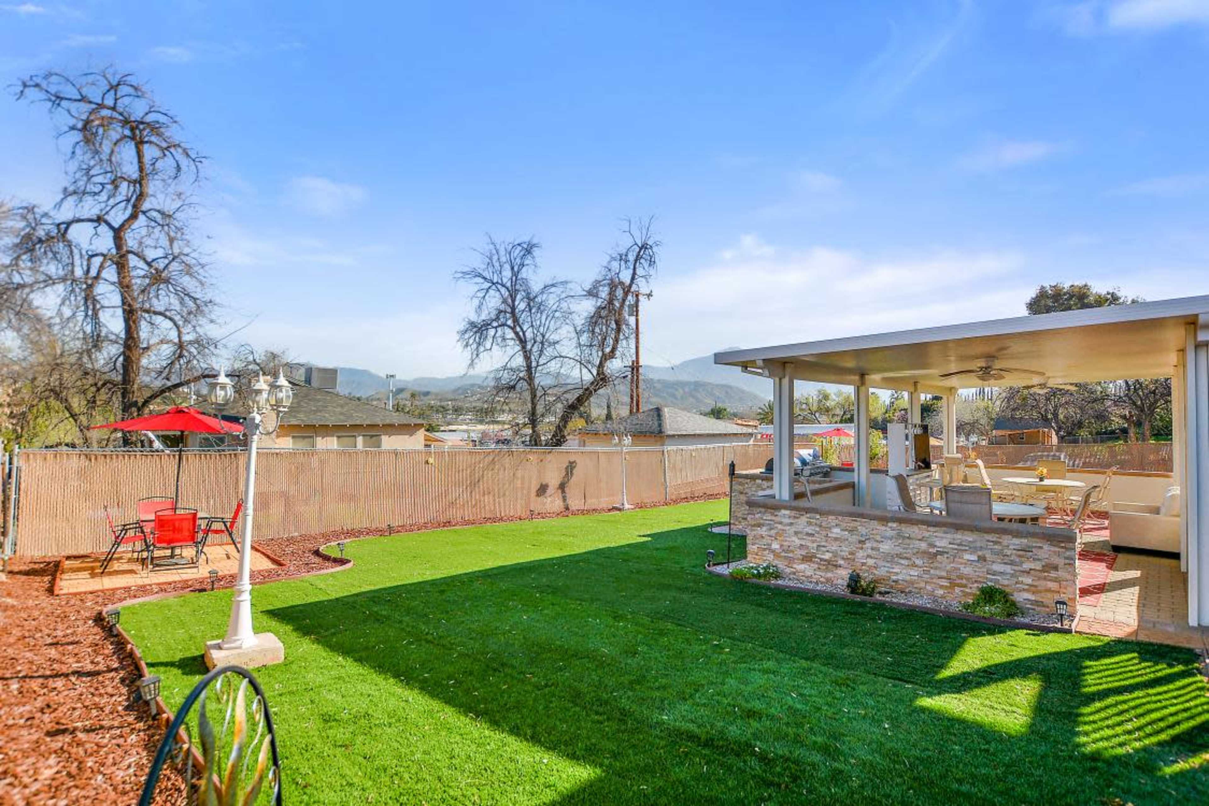 A backyard with a well-maintained lawn, a stone patio, a red umbrella over a table, and a fence surrounding the area, with mountains in the background.