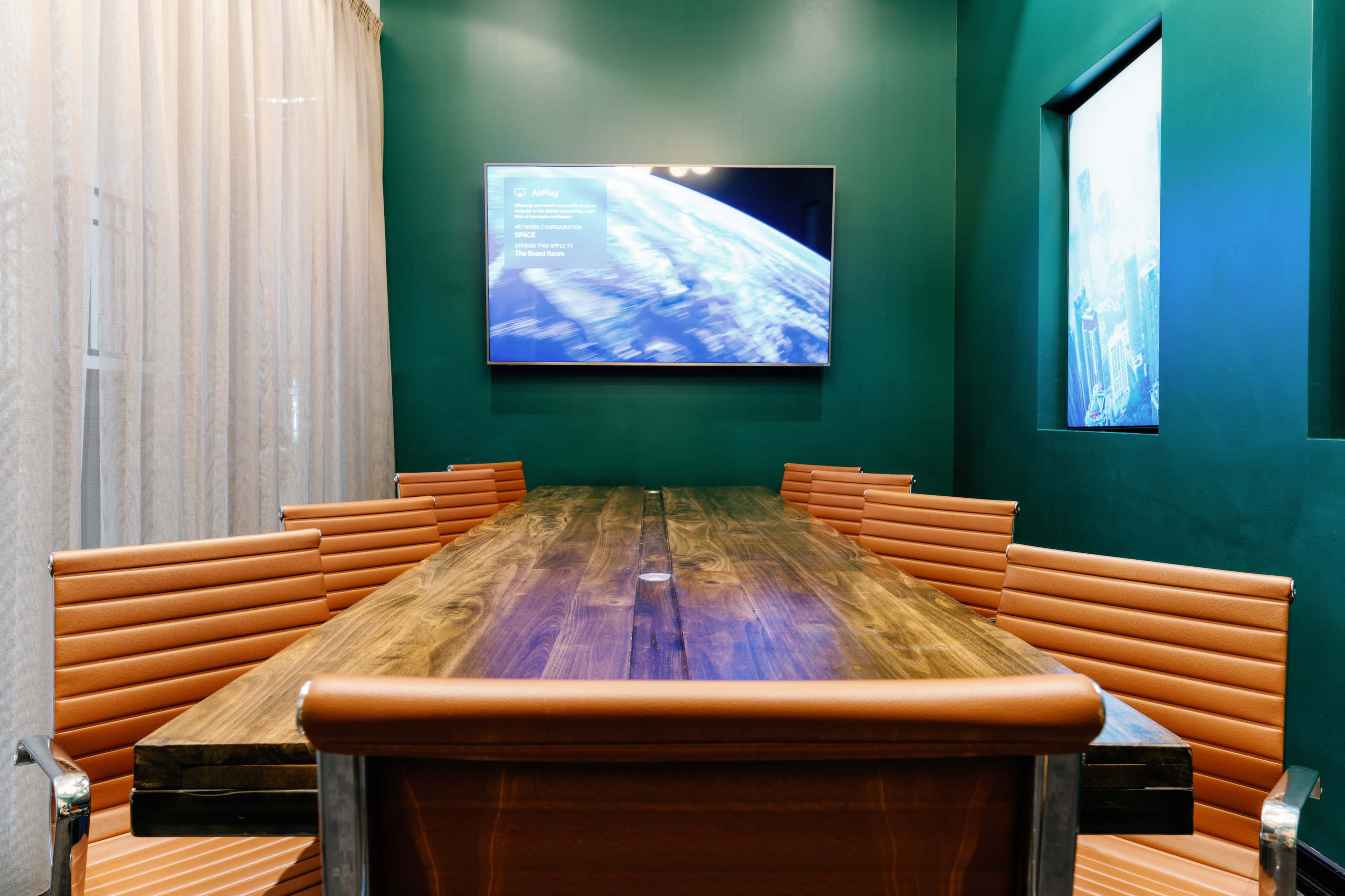The image shows a conference room featuring a long wooden table surrounded by orange striped chairs, with a television mounted on a green wall displaying information.