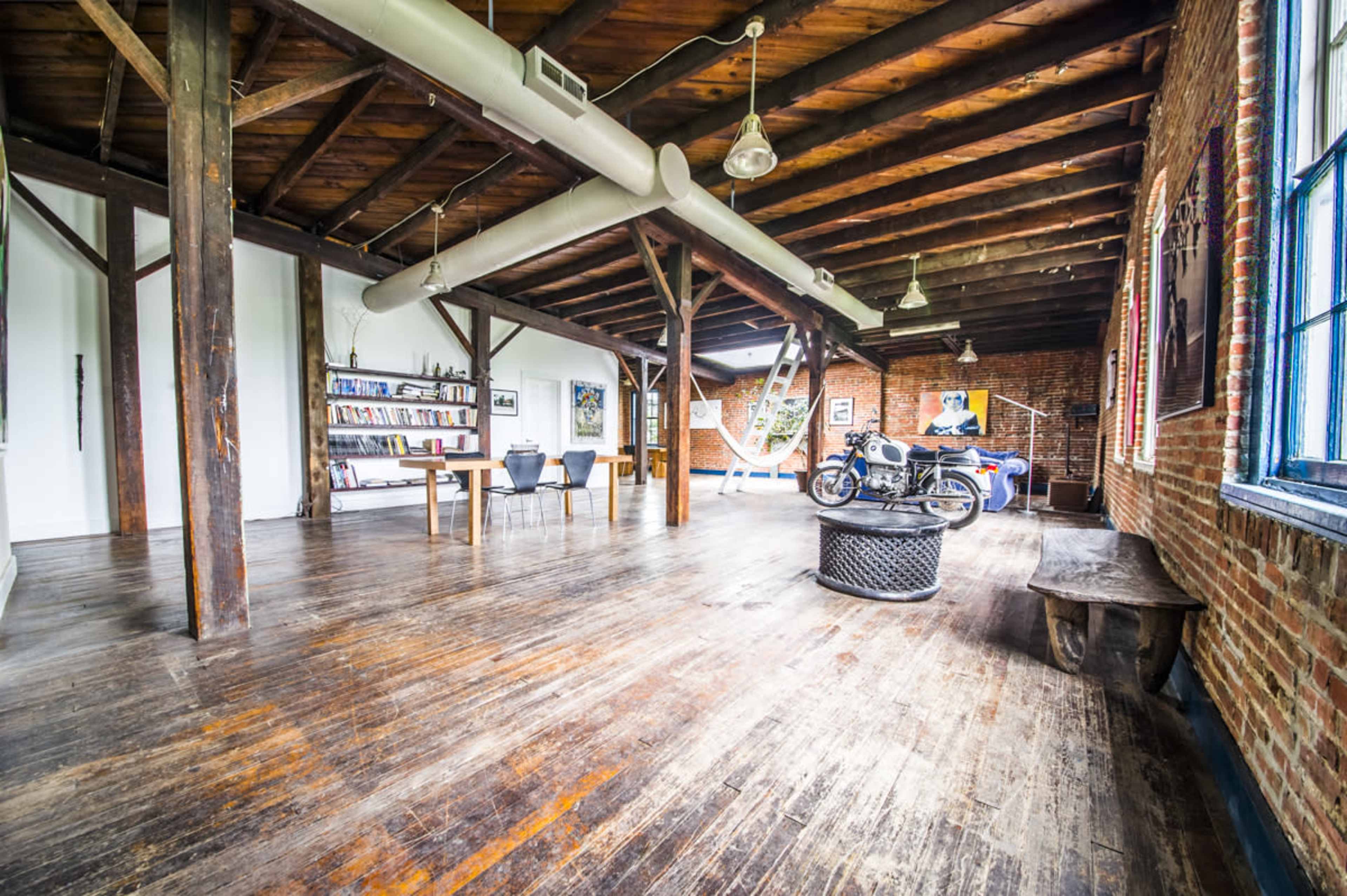 The image shows an open, spacious loft with wooden beams, a dining table, a motorcycle, and brick walls.