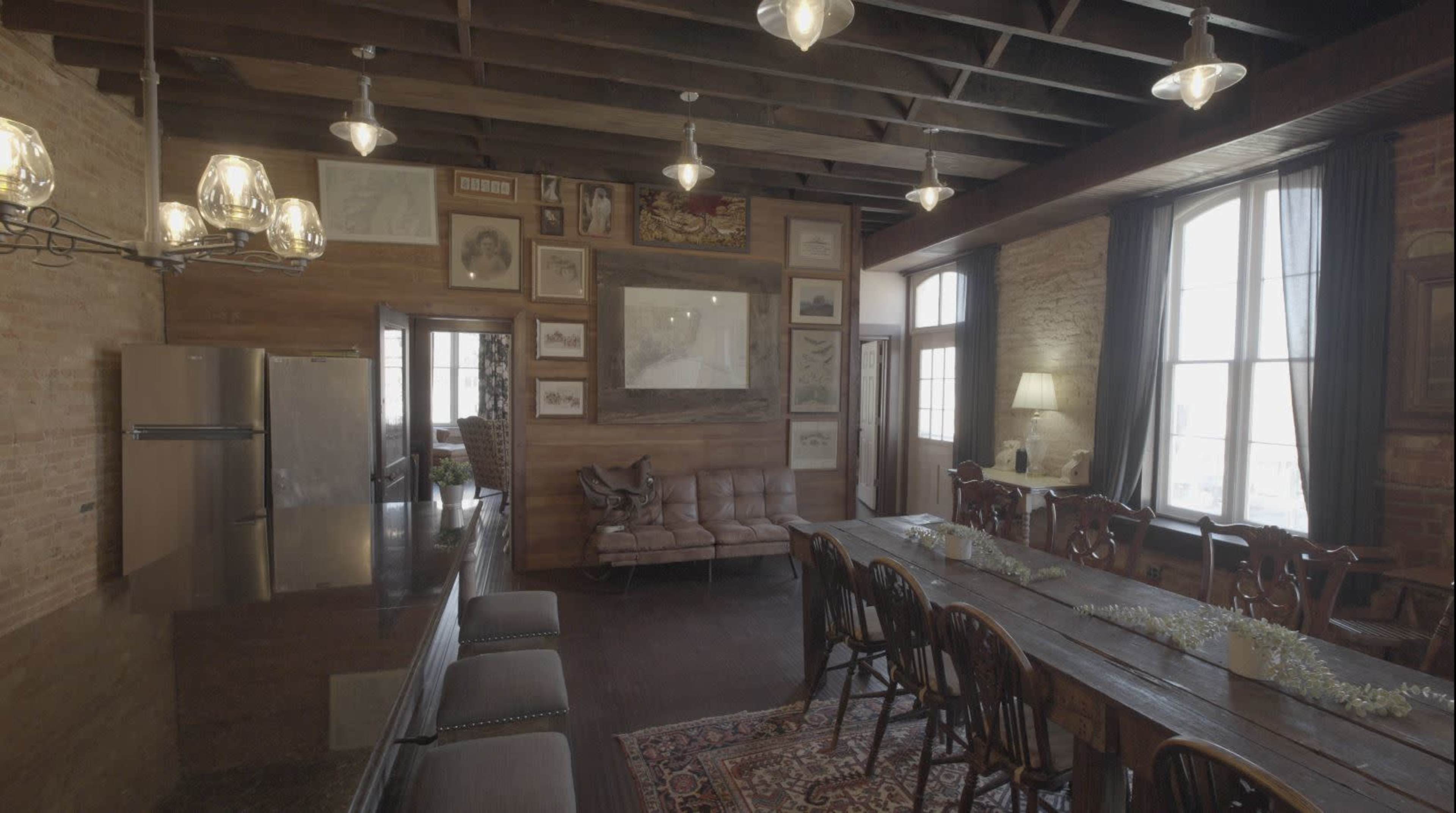 The image shows a spacious room with exposed brick walls, large windows, a long dining table, and a lounge area featuring a leather sofa.