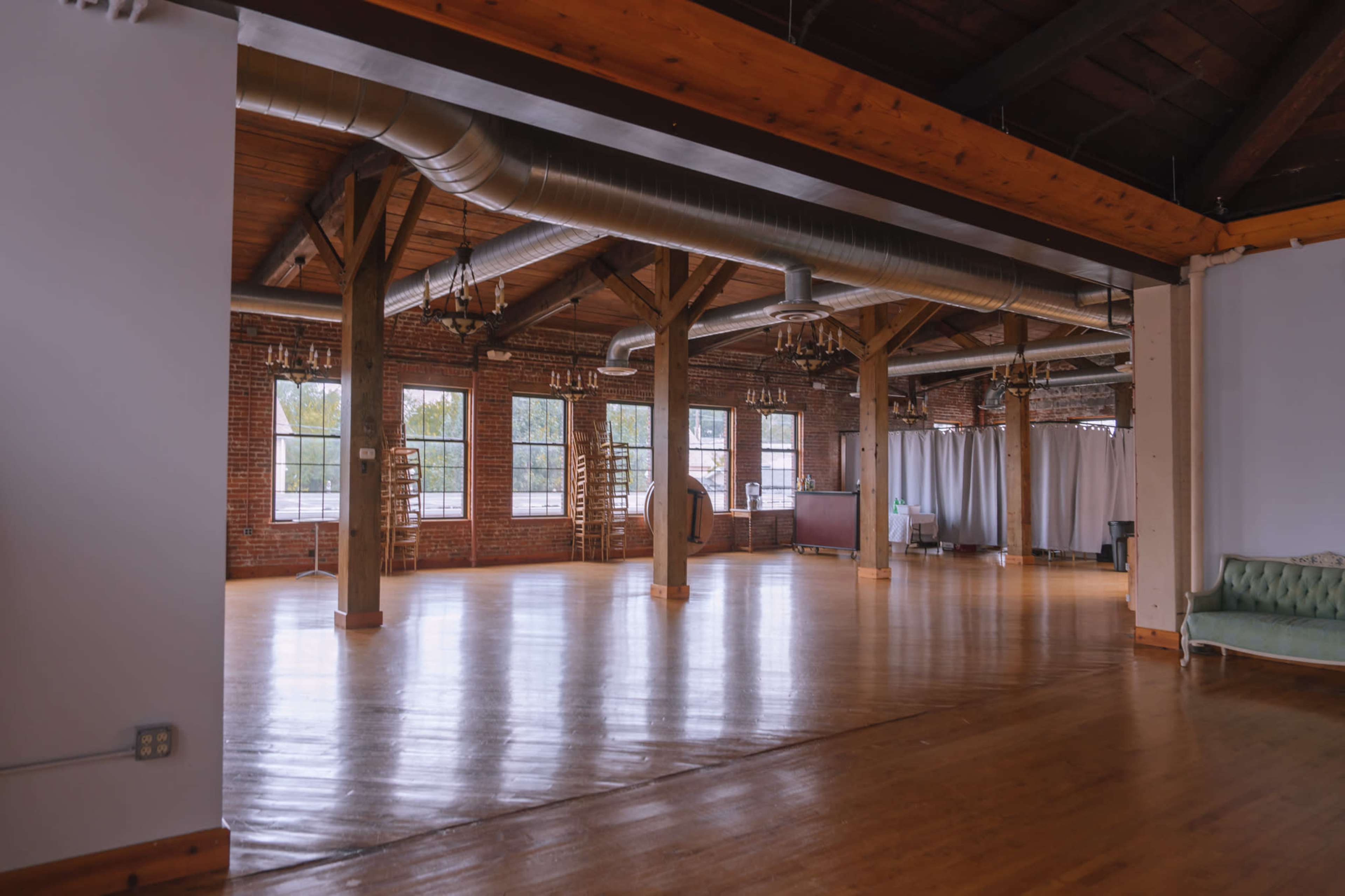The image shows a spacious, well-lit interior of a room with exposed wooden beams, large windows, and a polished wooden floor.