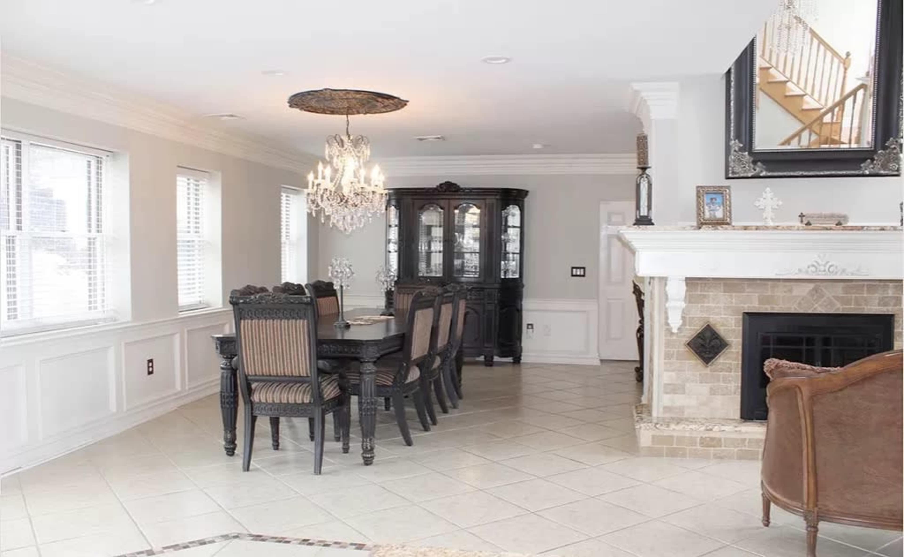 The image shows a dining area with a long table and eight chairs, a black cabinet, a chandelier, and a fireplace with a marble surround.