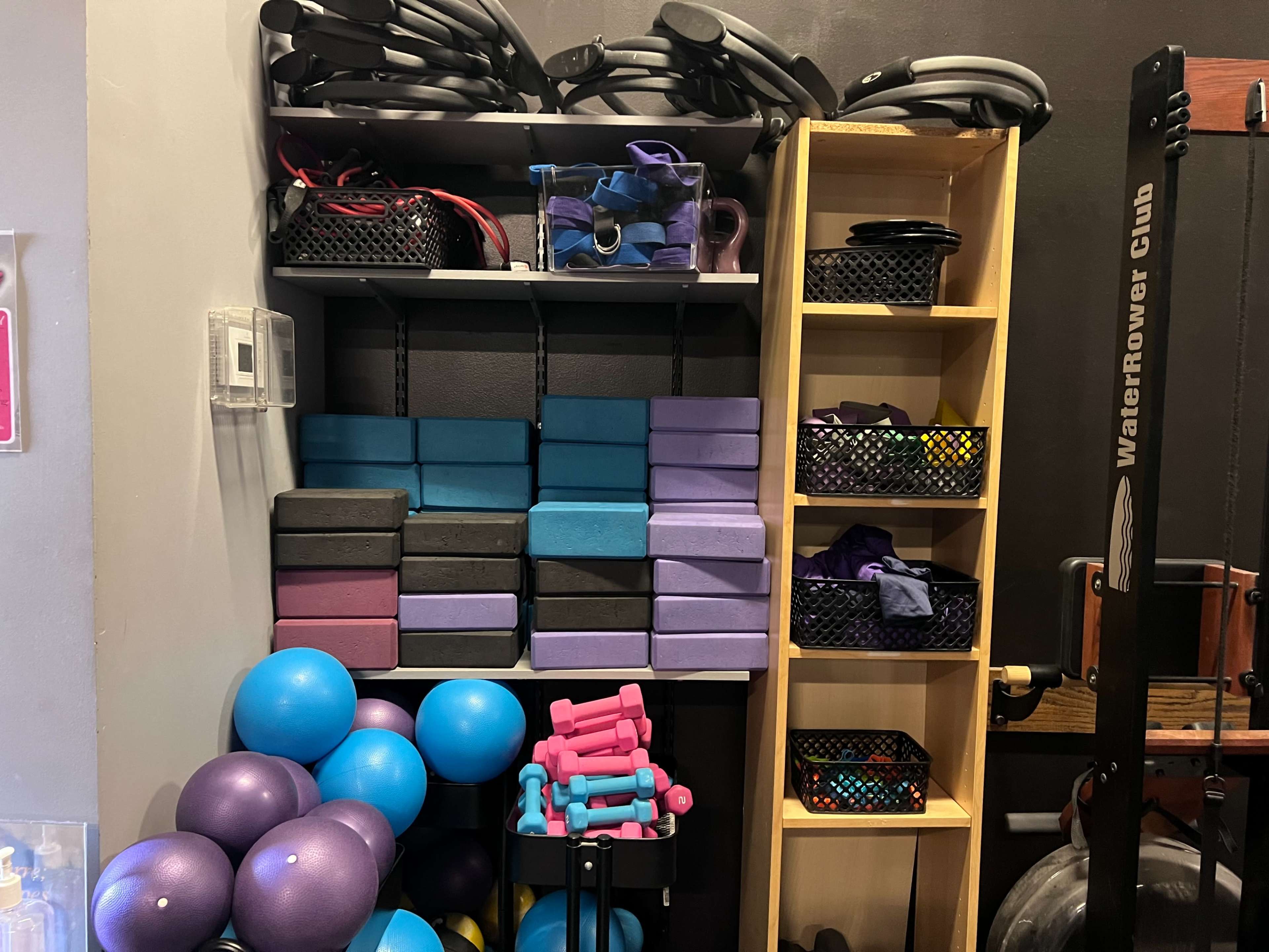 The image shows a fitness storage area with shelves containing various exercise equipment, including foam blocks, weights, and balls, organized in colored baskets.