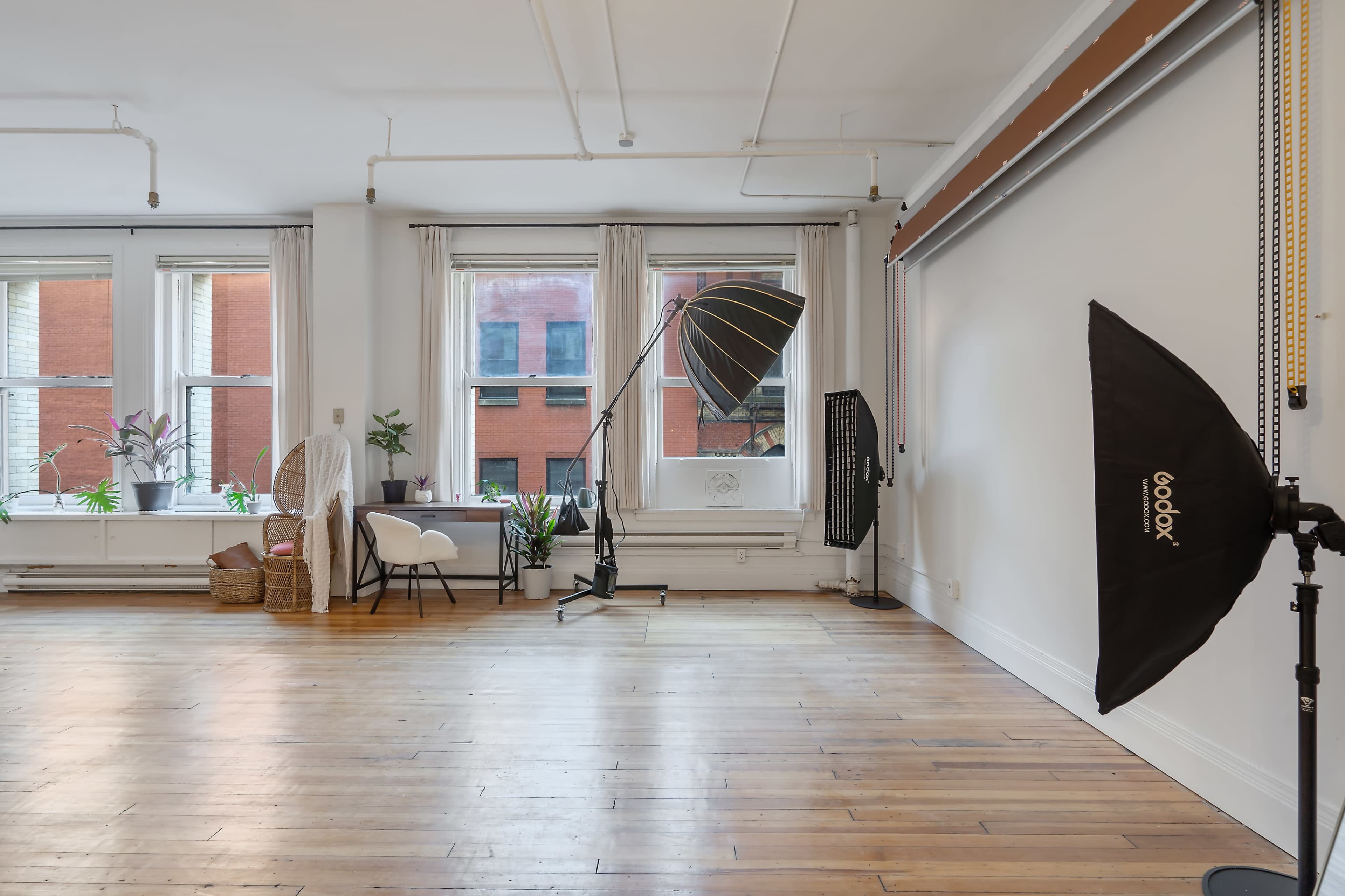A spacious, well-lit room features large windows, hardwood floors, and photography equipment, including a softbox and a backdrop stand.