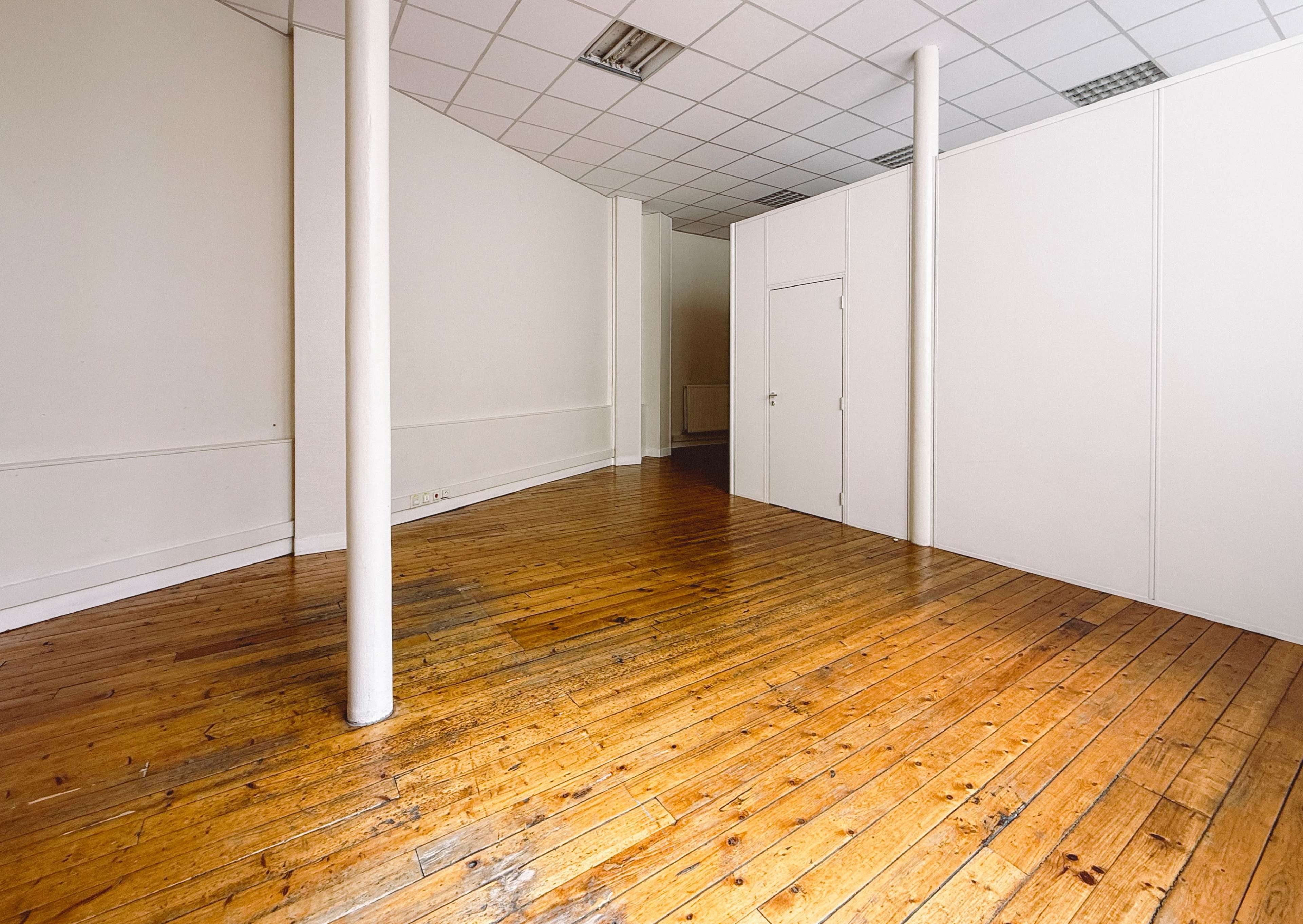 The image shows an empty room with wooden flooring, white walls, and two columns.