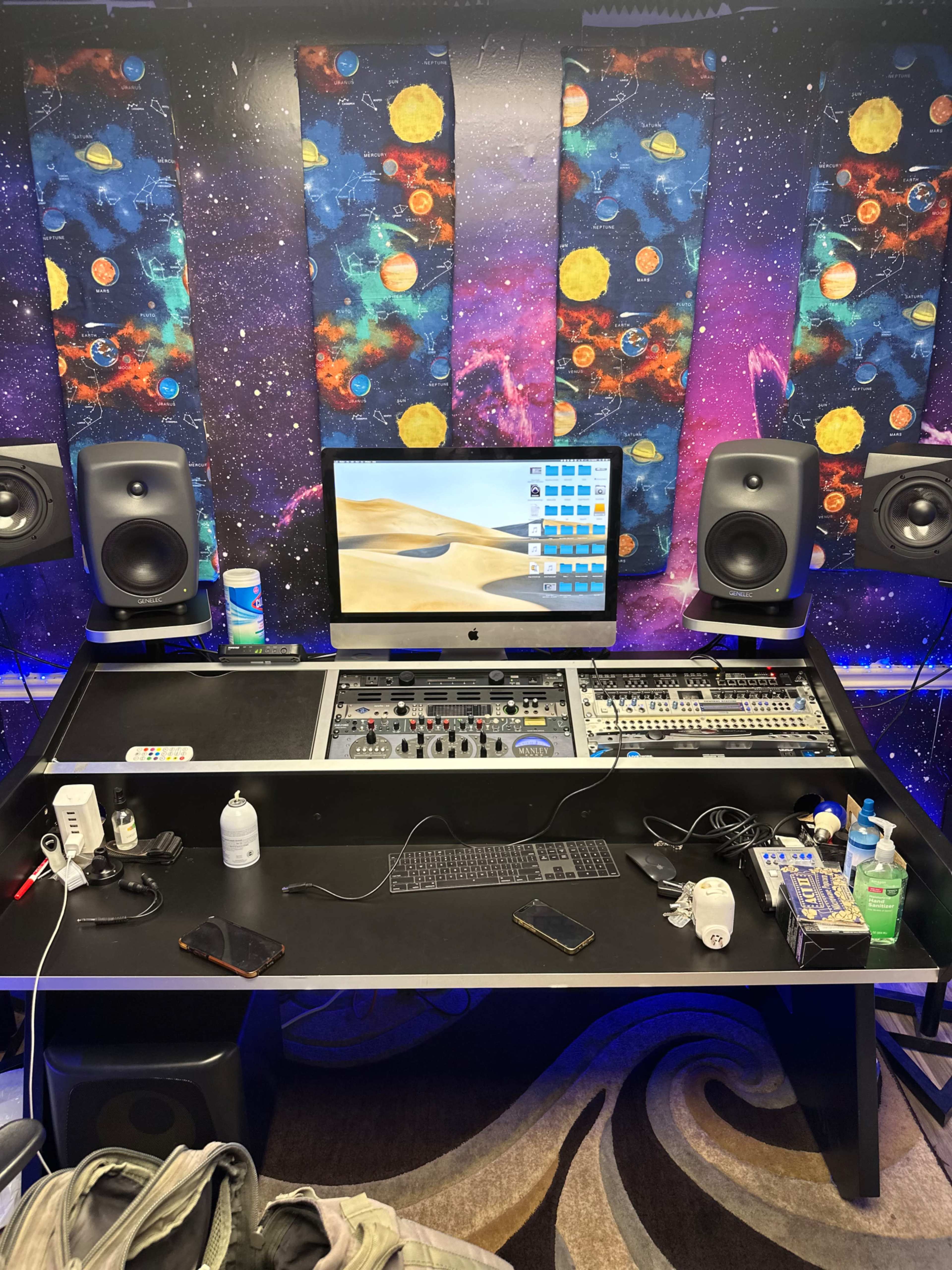 A recording studio featuring a large desk with audio equipment, speakers, a computer, and various personal items on the surface, set against a colorful cosmic-themed backdrop.