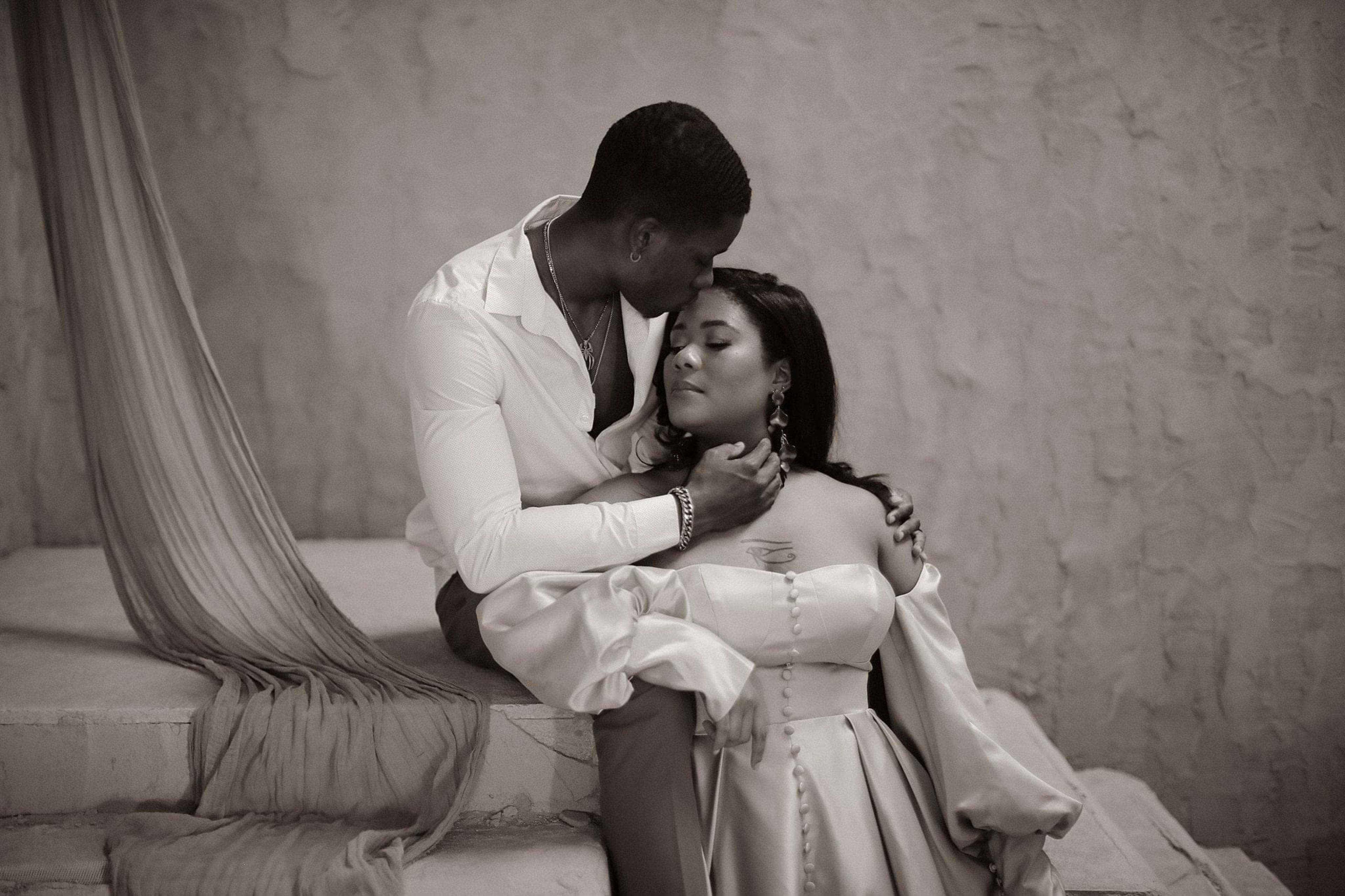 A couple during a black and white photoshoot, with the man kneeling down next to the woman in a dress.