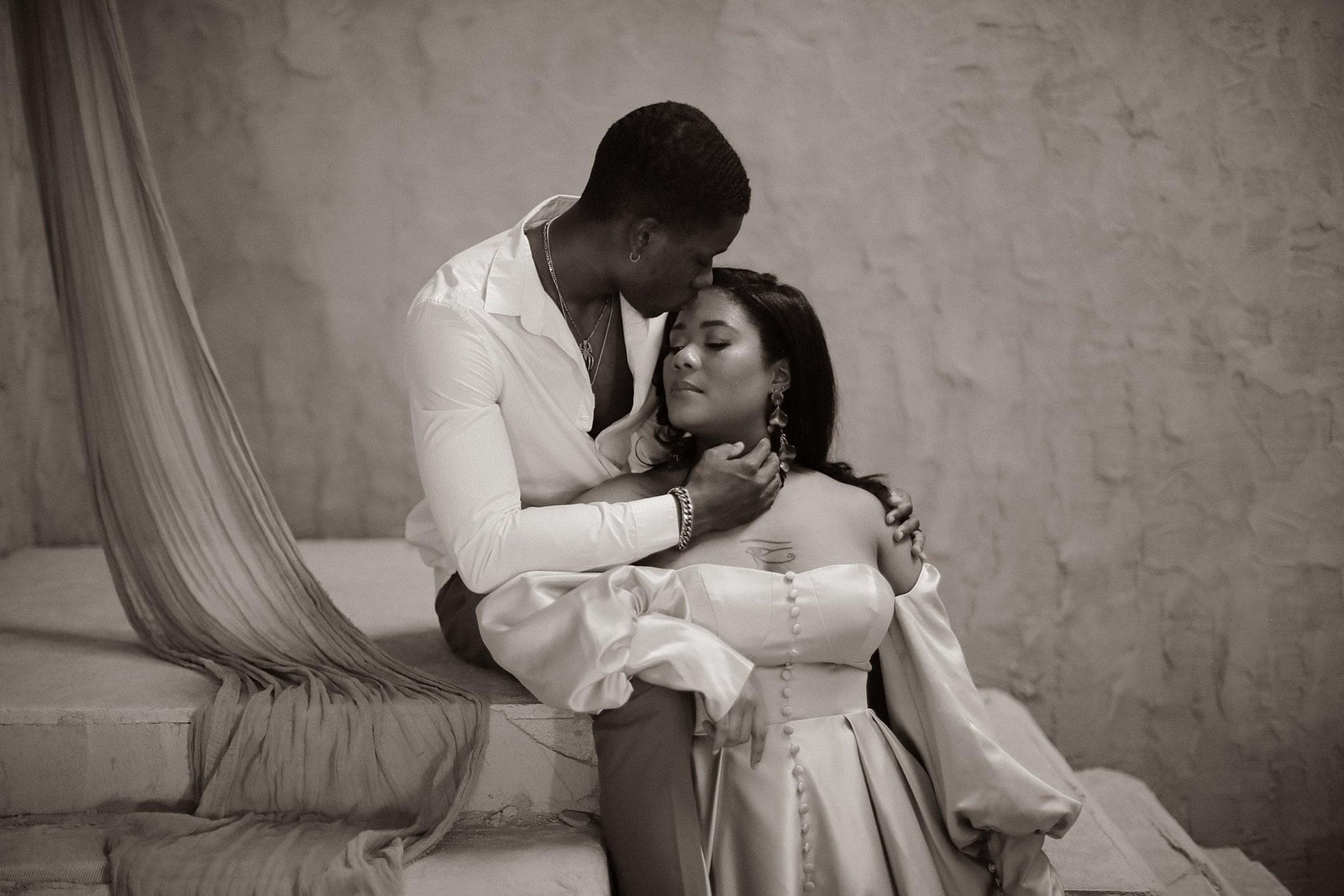 Una pareja durante una sesión de fotos en blanco y negro, con el hombre arrodillado junto a la mujer ataviada con un vestido.