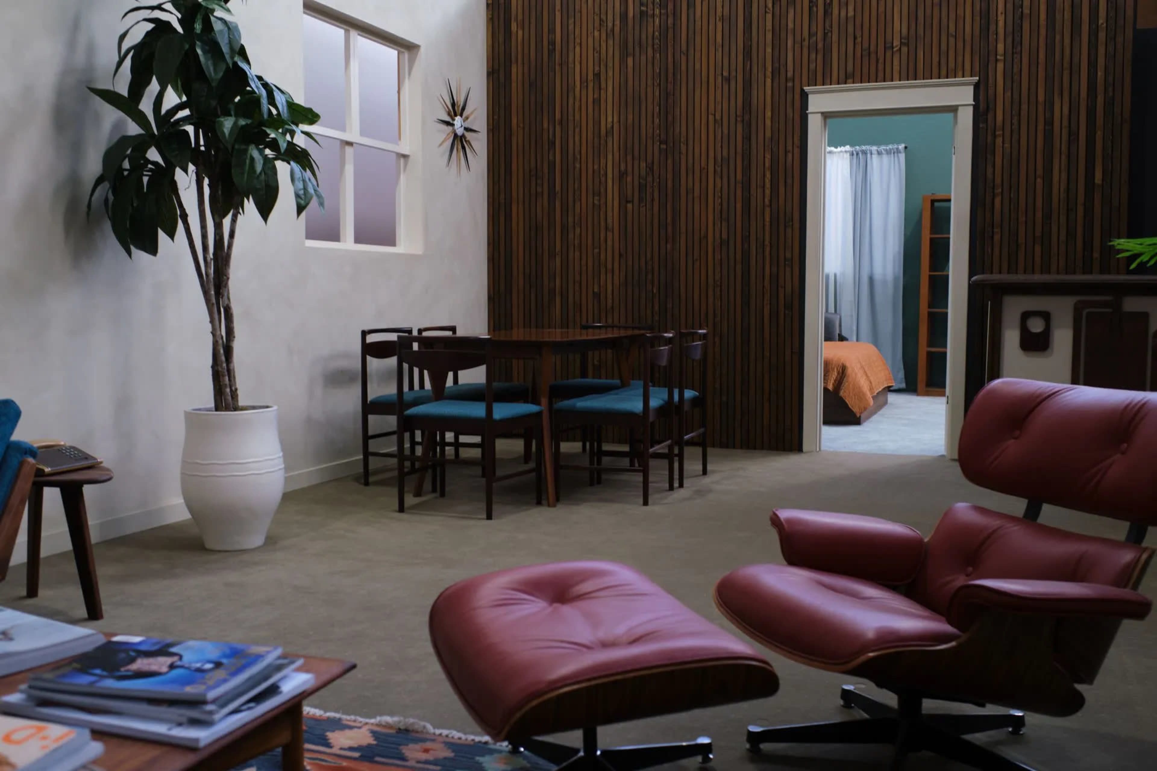 The image shows a modern interior design featuring a brown wooden wall, a dining area with a circular table and teal chairs, and a seating area with red lounge chairs.