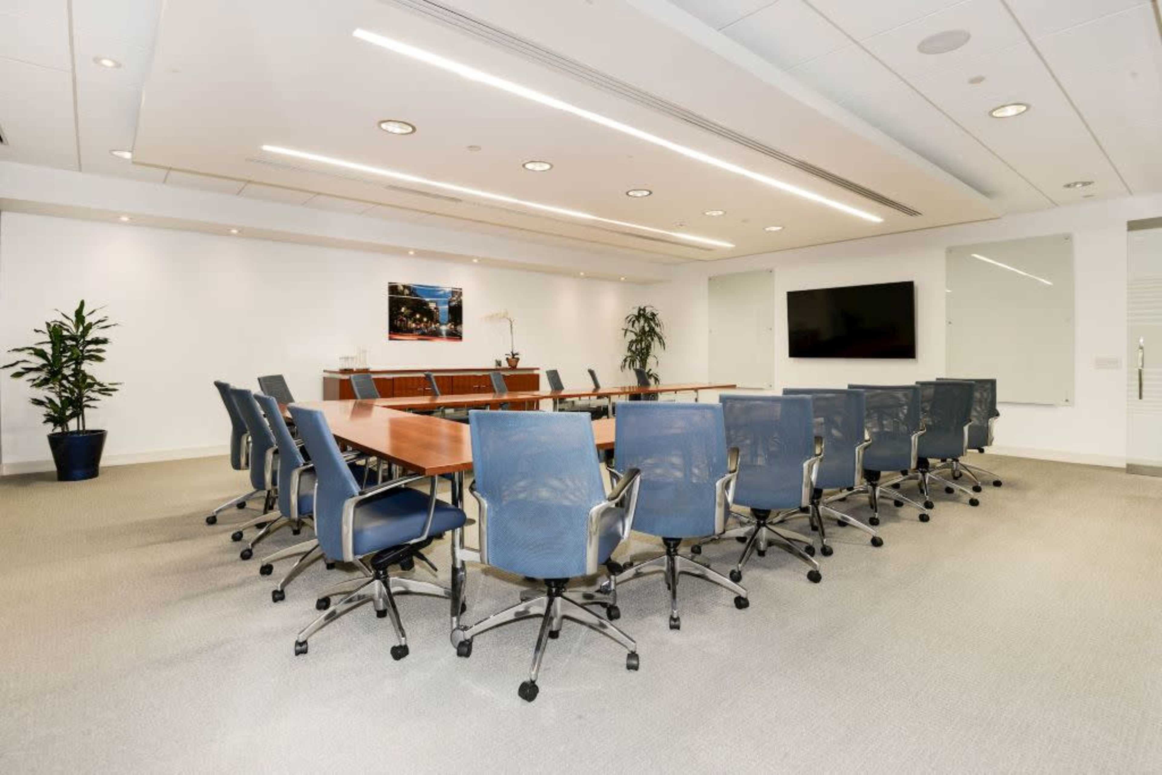 A modern conference room features a long rectangular table surrounded by ergonomic chairs, with a large screen and plants in the background.