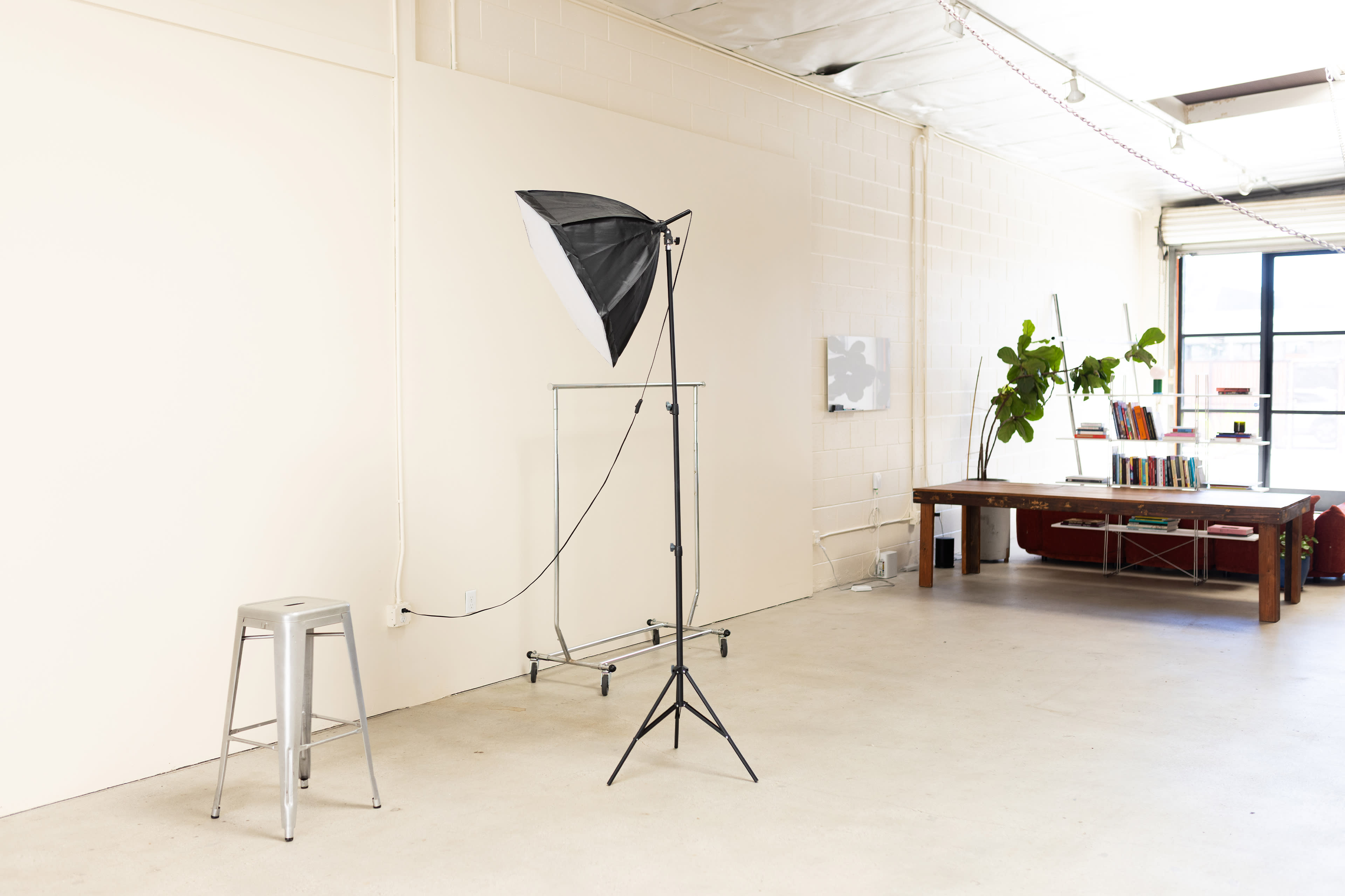 The image shows a bright, minimalistic studio space featuring a photography light on a stand next to a metal stool, with a wooden table and shelving in the background.