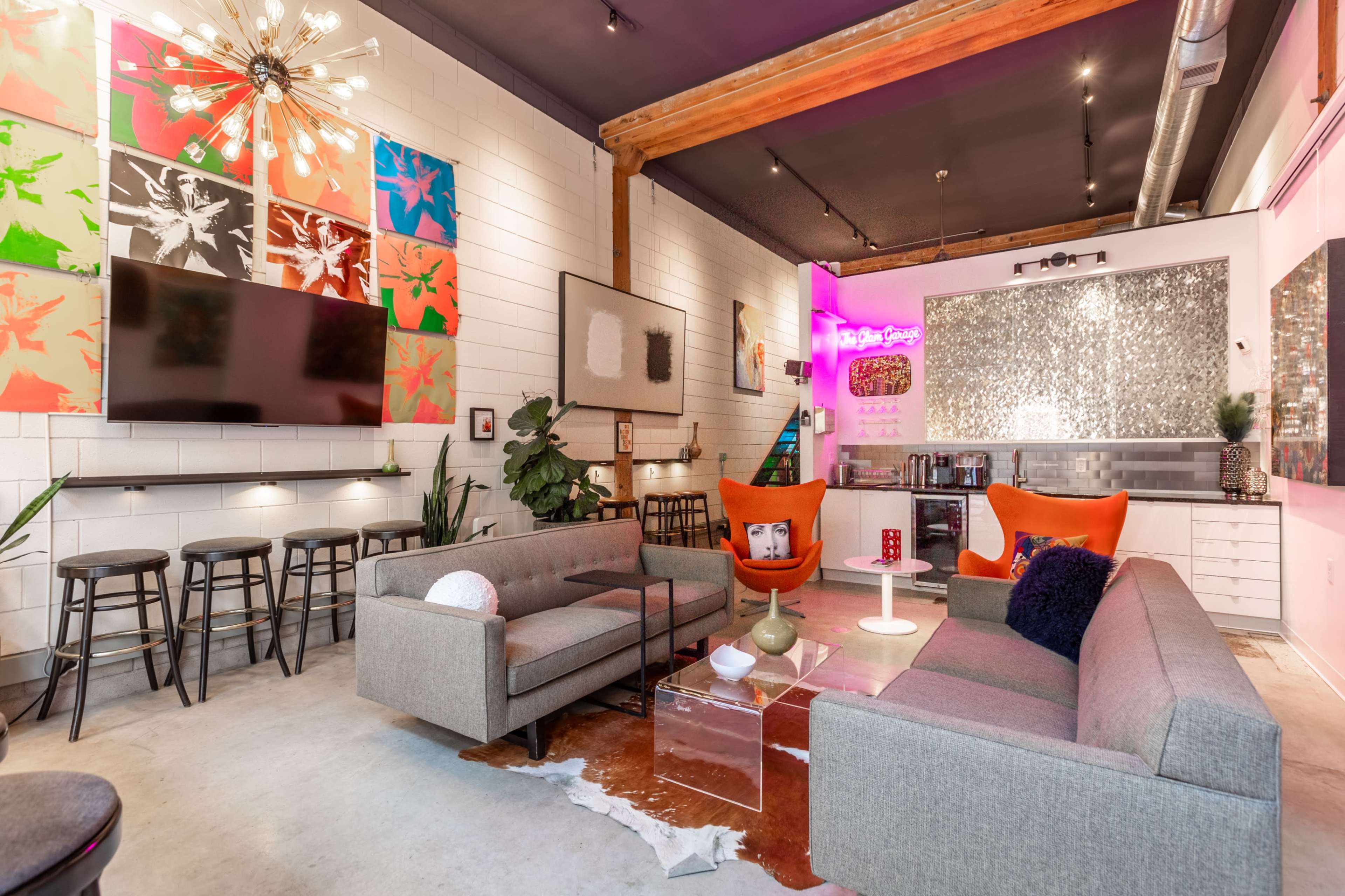 The image features a stylish modern living space with gray sofas, vibrant orange accent chairs, and artwork on the walls, complemented by a bar area with high stools and a television.