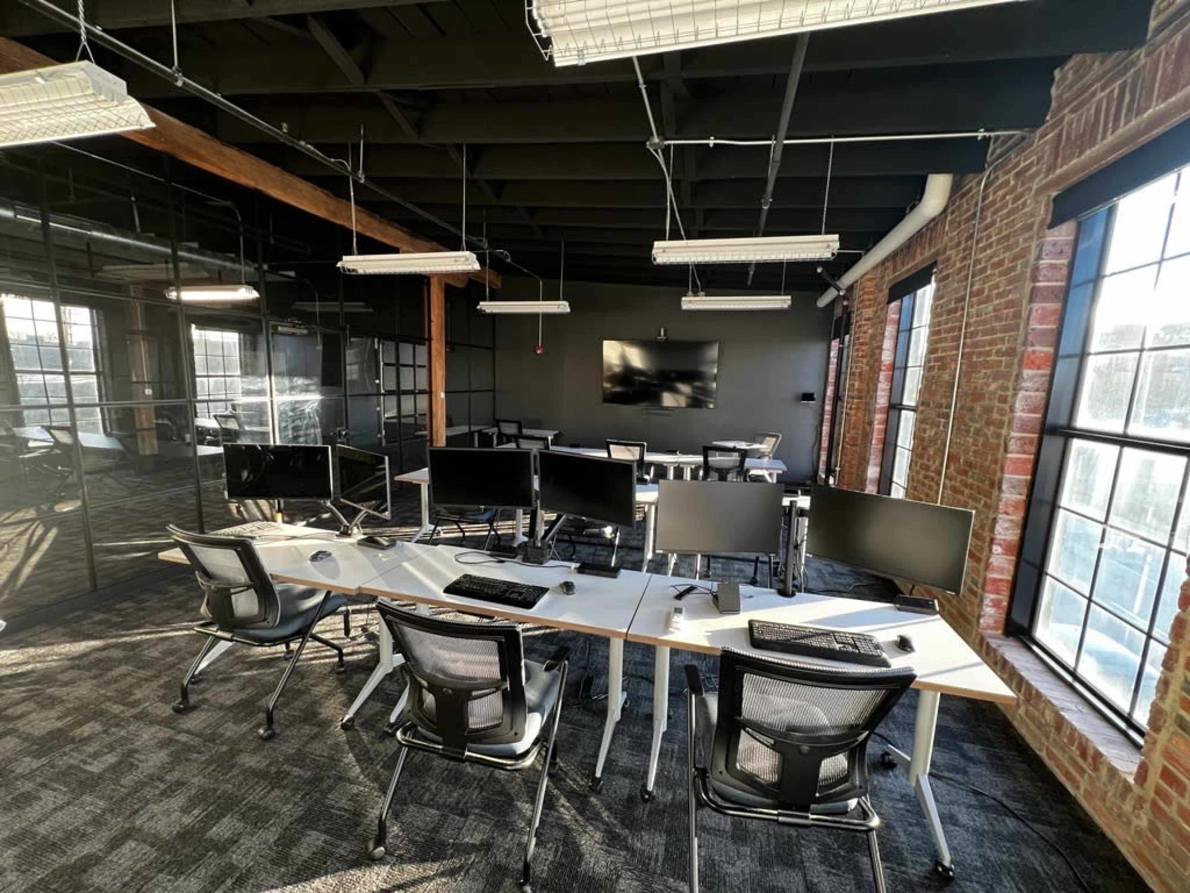 A modern office space featuring multiple workstations with computer monitors and a conference area with a large screen, surrounded by large windows and exposed brick walls.