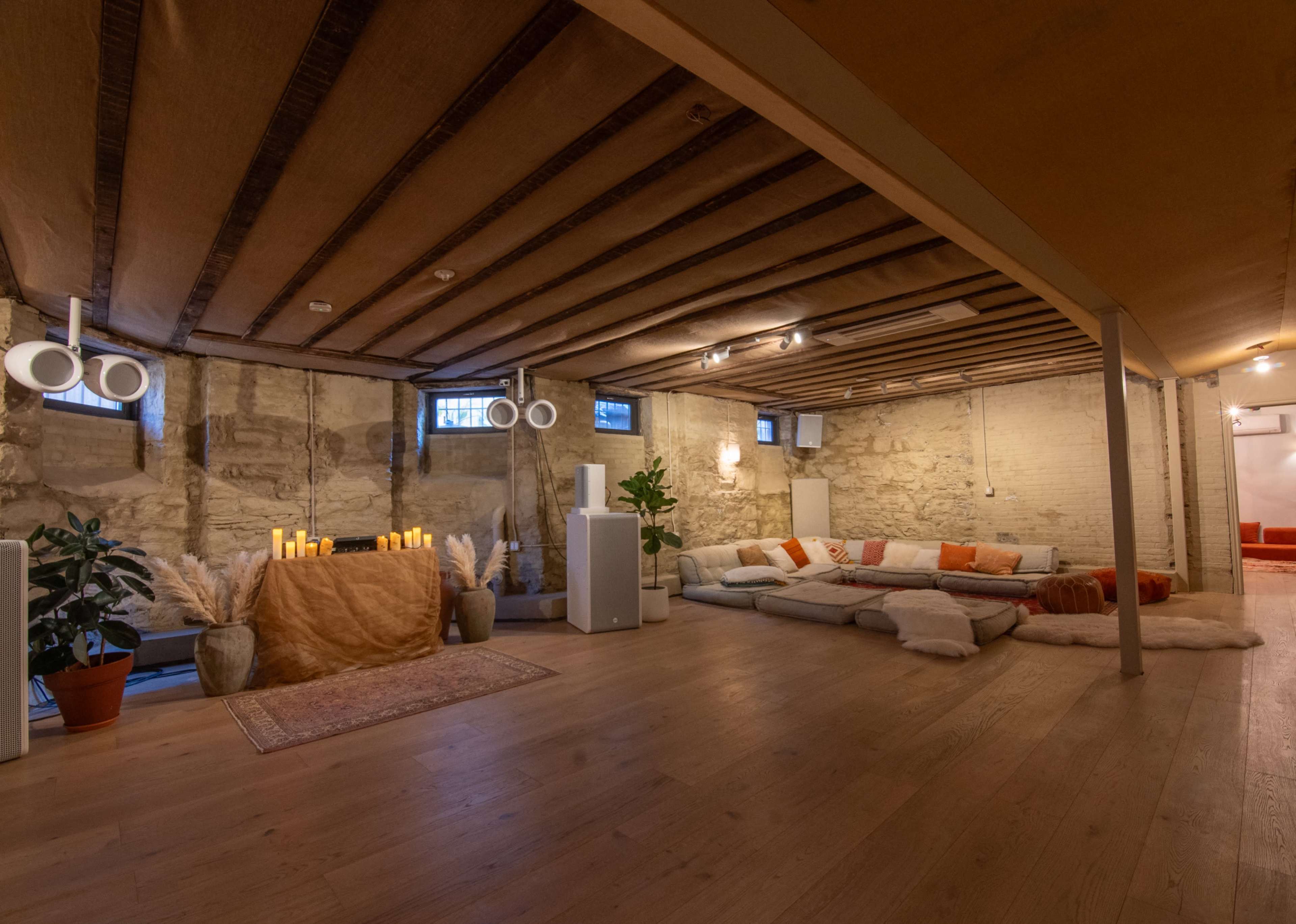 The image shows a spacious, well-lit basement area featuring a cozy seating arrangement with cushions, a decorative table with candles, and potted plants.