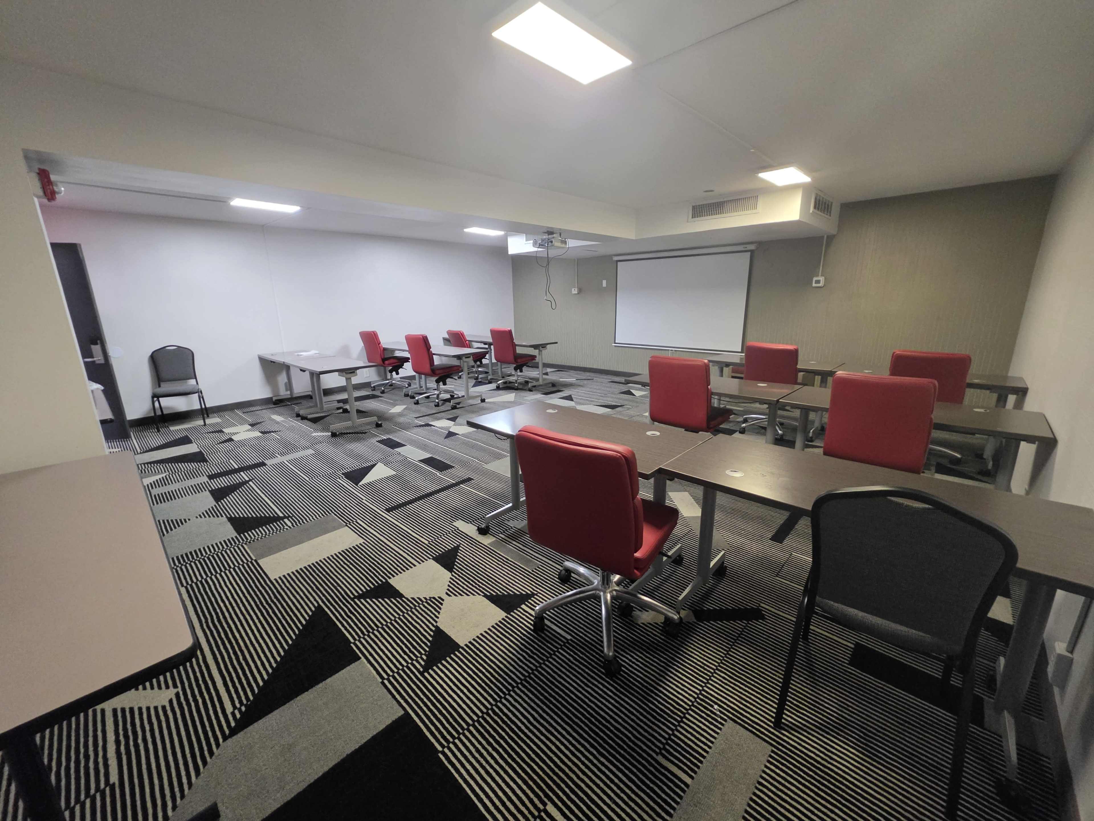 A conference room features several tables and red chairs arranged on a patterned carpet, with a projector screen mounted on the wall.