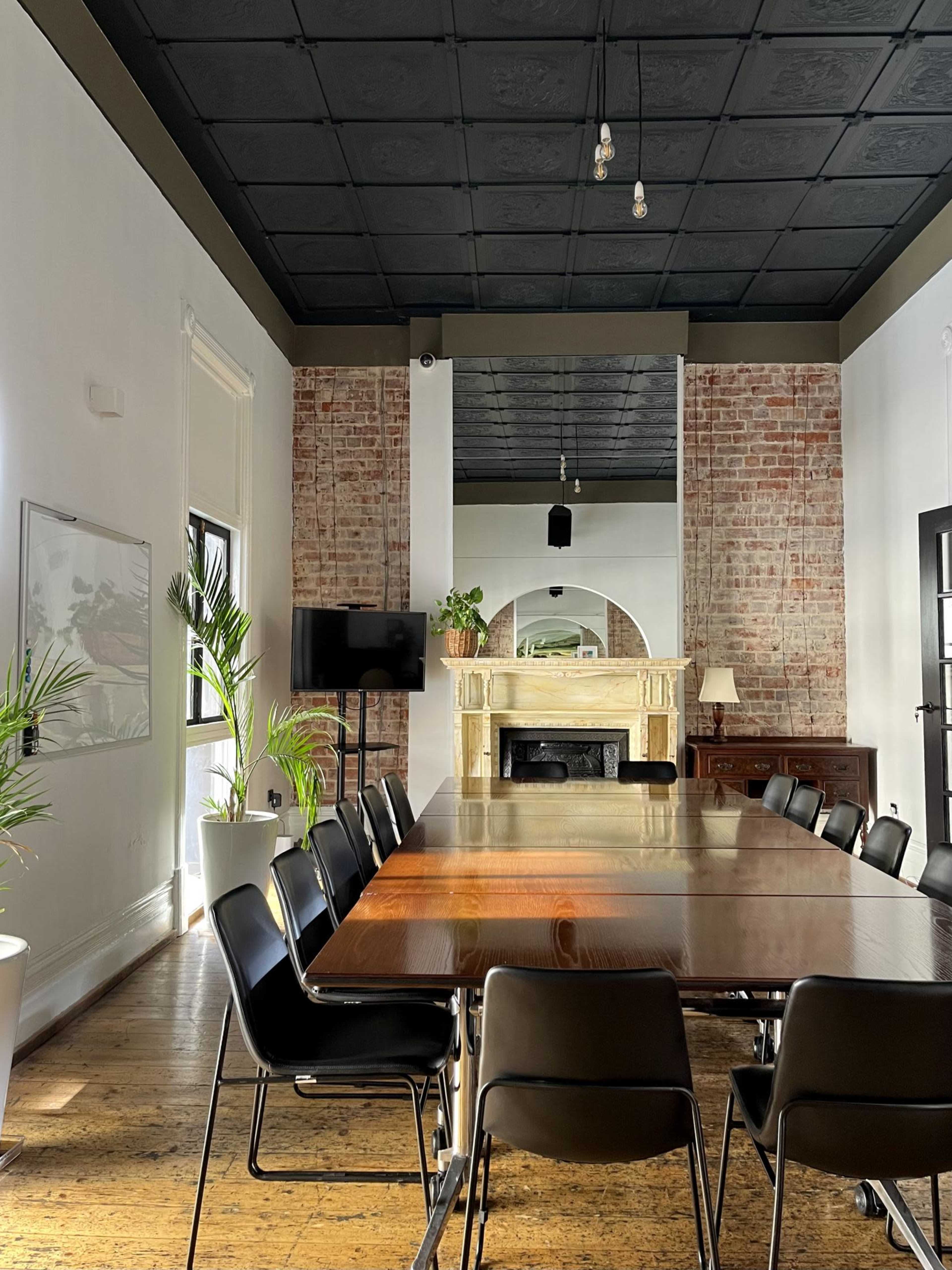 A long wooden table surrounded by black chairs is set in a spacious room with exposed brick walls, a fireplace, and large windows allowing natural light to enter.