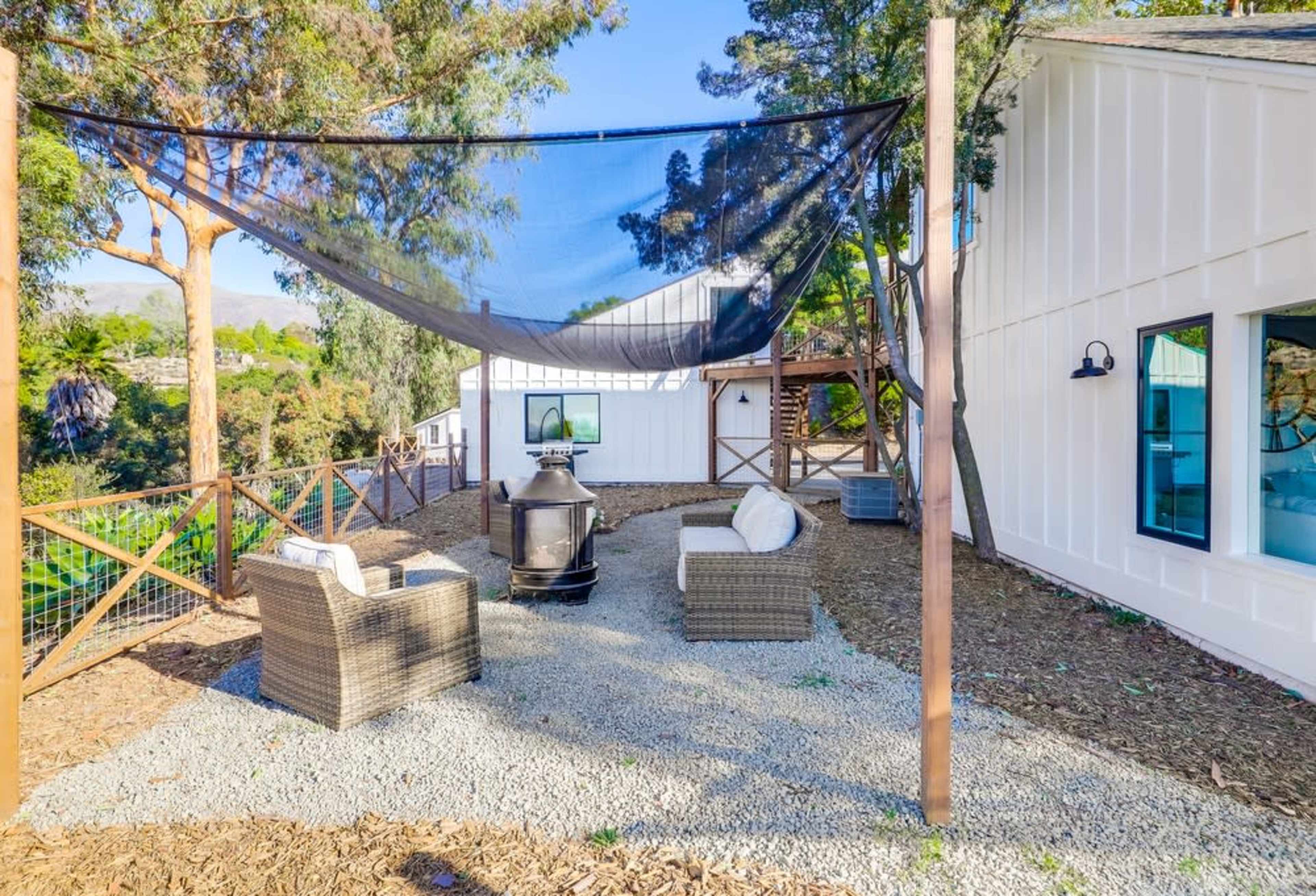 A shaded patio area with two wicker chairs, a fire pit, and a backdrop of trees and a white building.