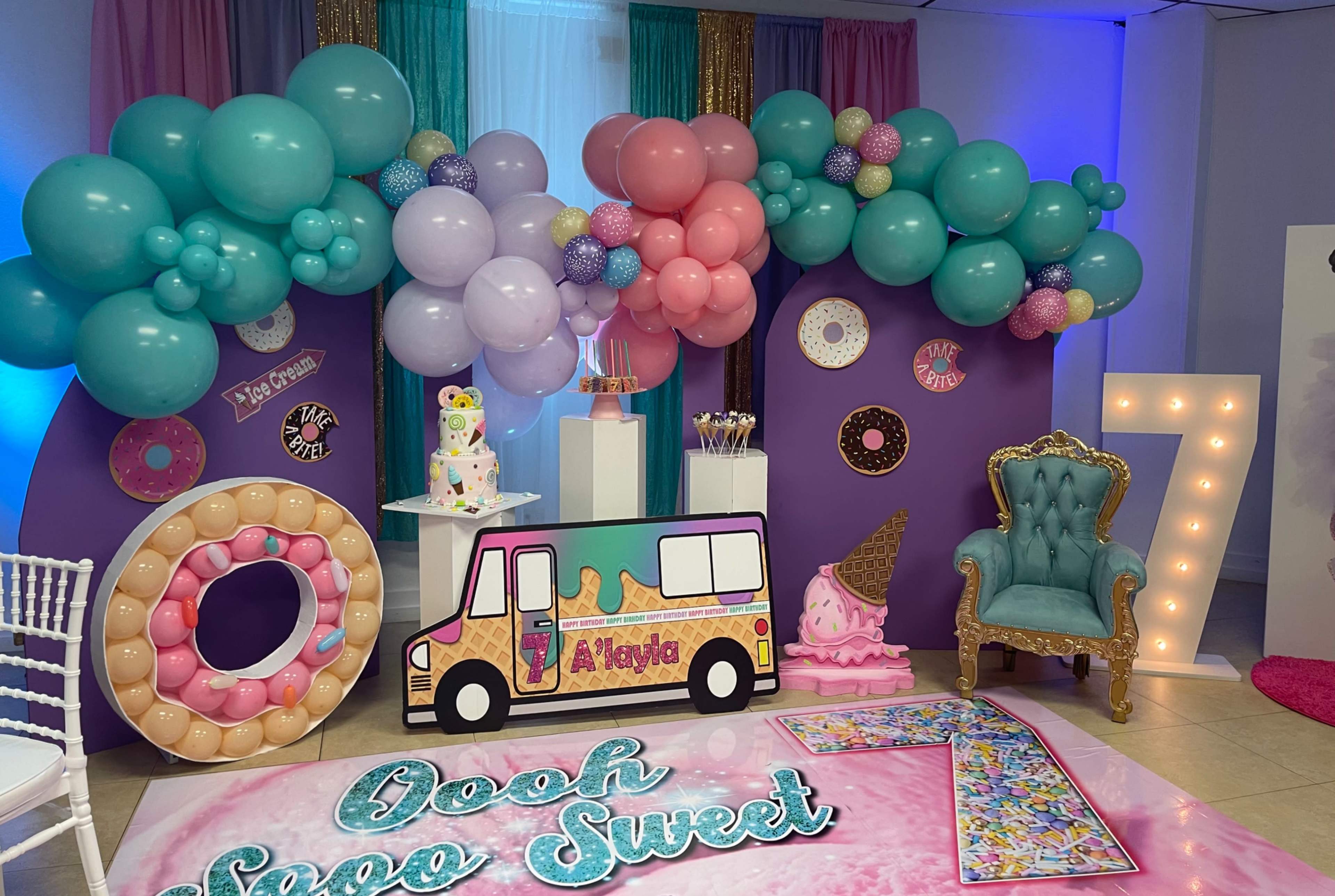 The image depicts a colorful birthday party setup featuring a decorative backdrop with balloons, a dessert table, and large cutouts of donuts and the number seven.