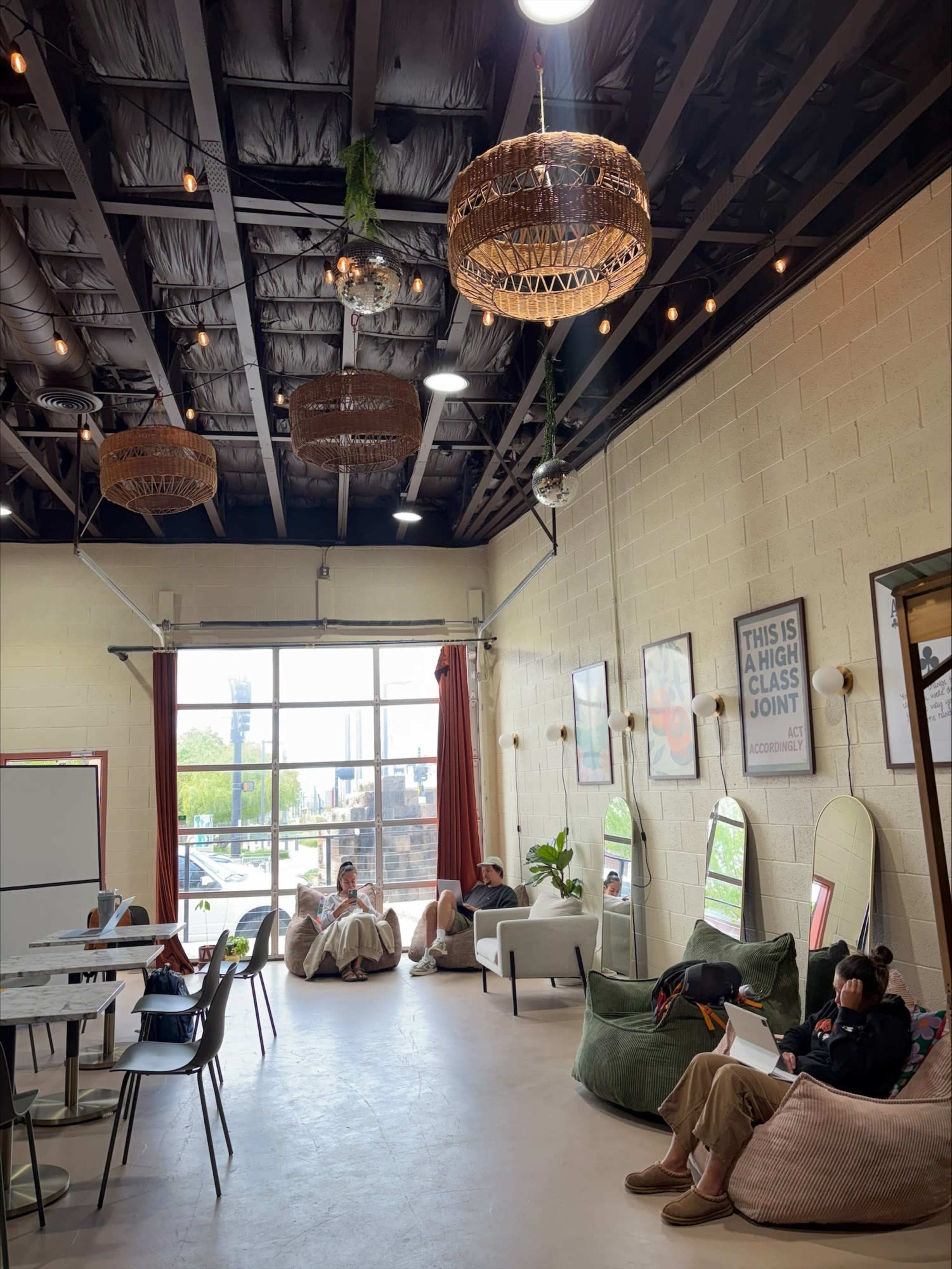 The image shows a spacious, modern interior of a lounge with people seated, plants, and unique overhead lighting fixtures.