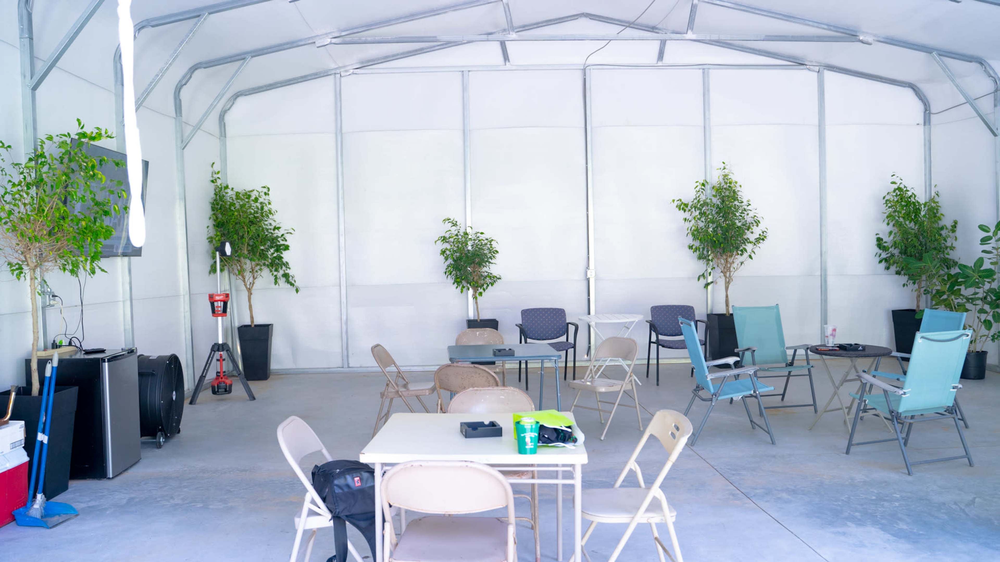 An indoor space with several tables and chairs arranged around potted plants, with a blank wall and a television visible in the background.