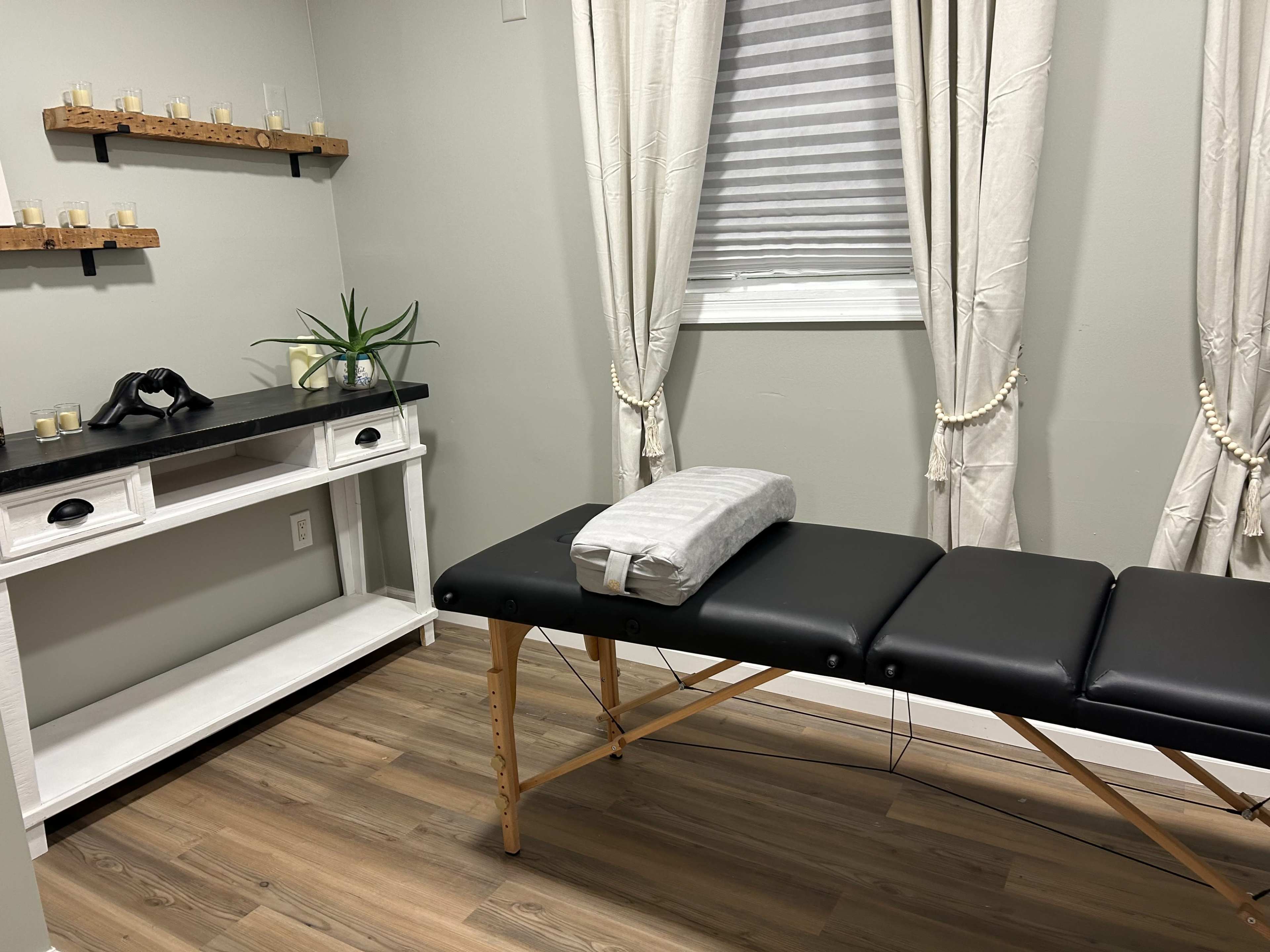 A small, well-organized treatment room featuring a black massage table, a white table with decorative items, and natural light filtering through sheer curtains.