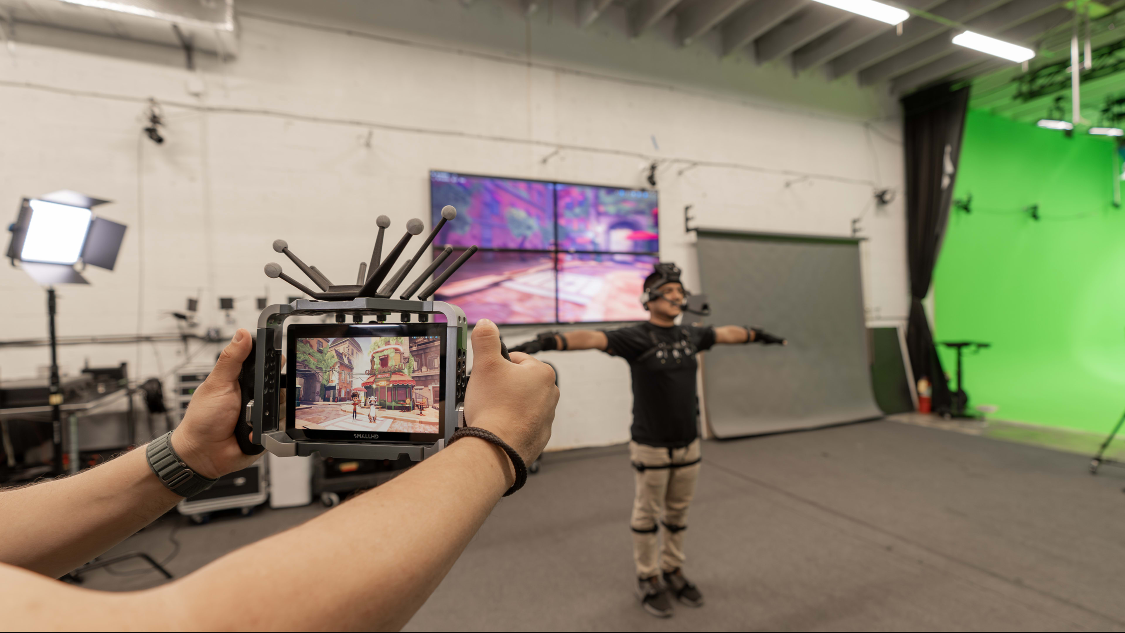 A person wearing motion capture gear stands in a studio while another person holds a tablet displaying a virtual environment.
