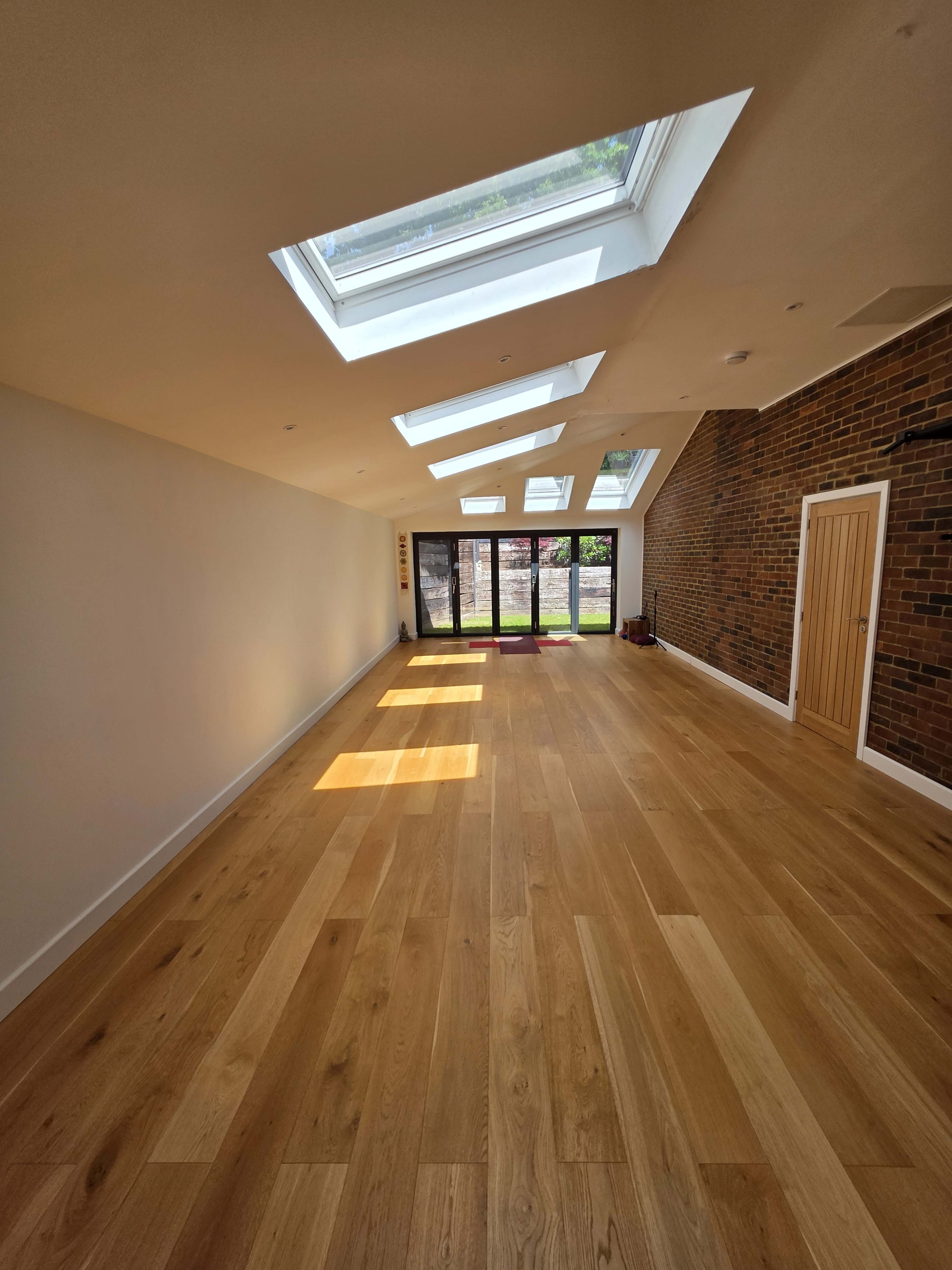 The image shows a spacious room with wooden flooring, skylights, and large glass doors leading to an outdoor area.