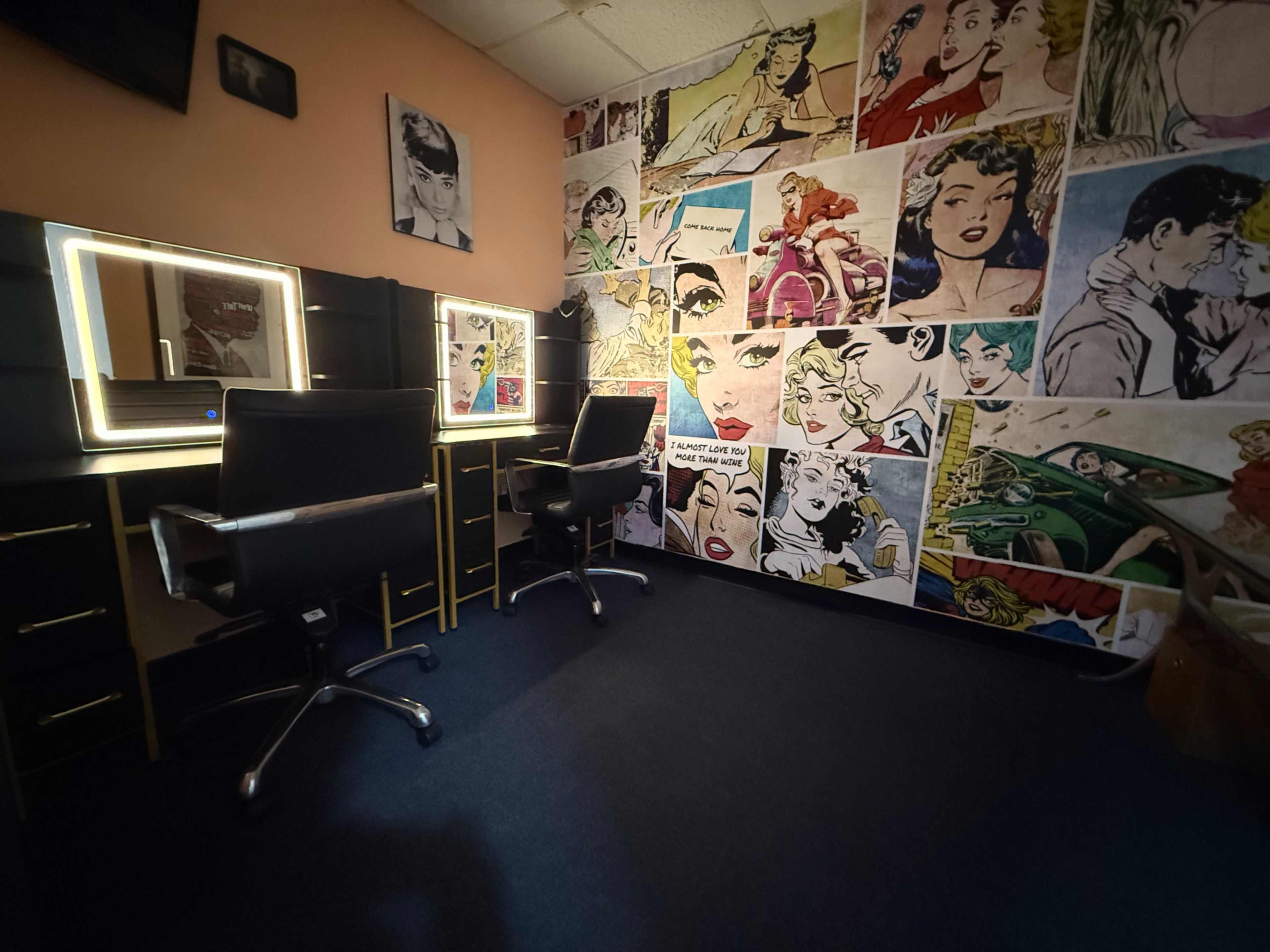 The image shows a room with two makeup stations illuminated by backlit mirrors, surrounded by a wall covered in colorful vintage comic book art.