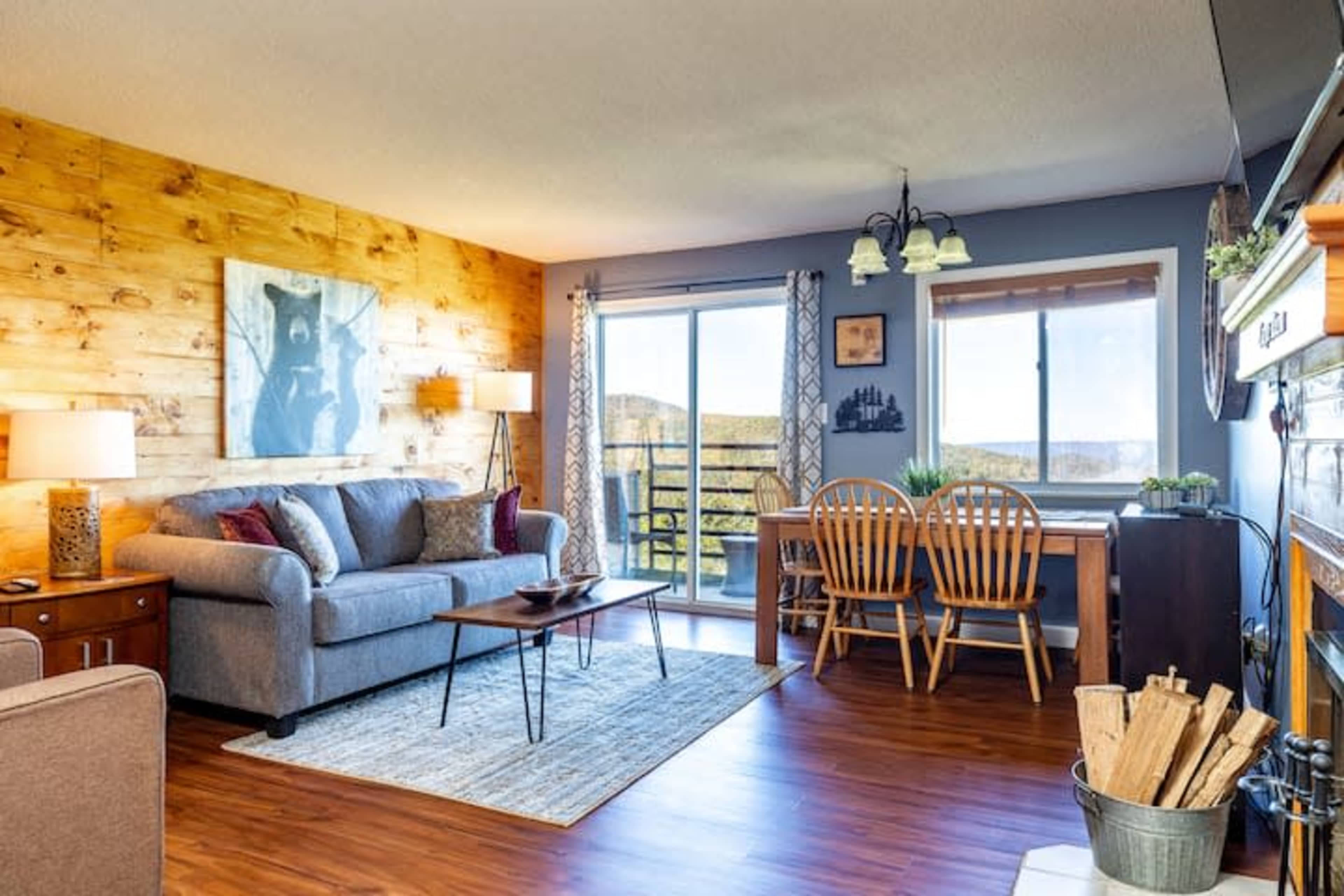 A cozy living area features a gray sofa, a wooden dining table with chairs, and large windows showcasing a view of the mountains.