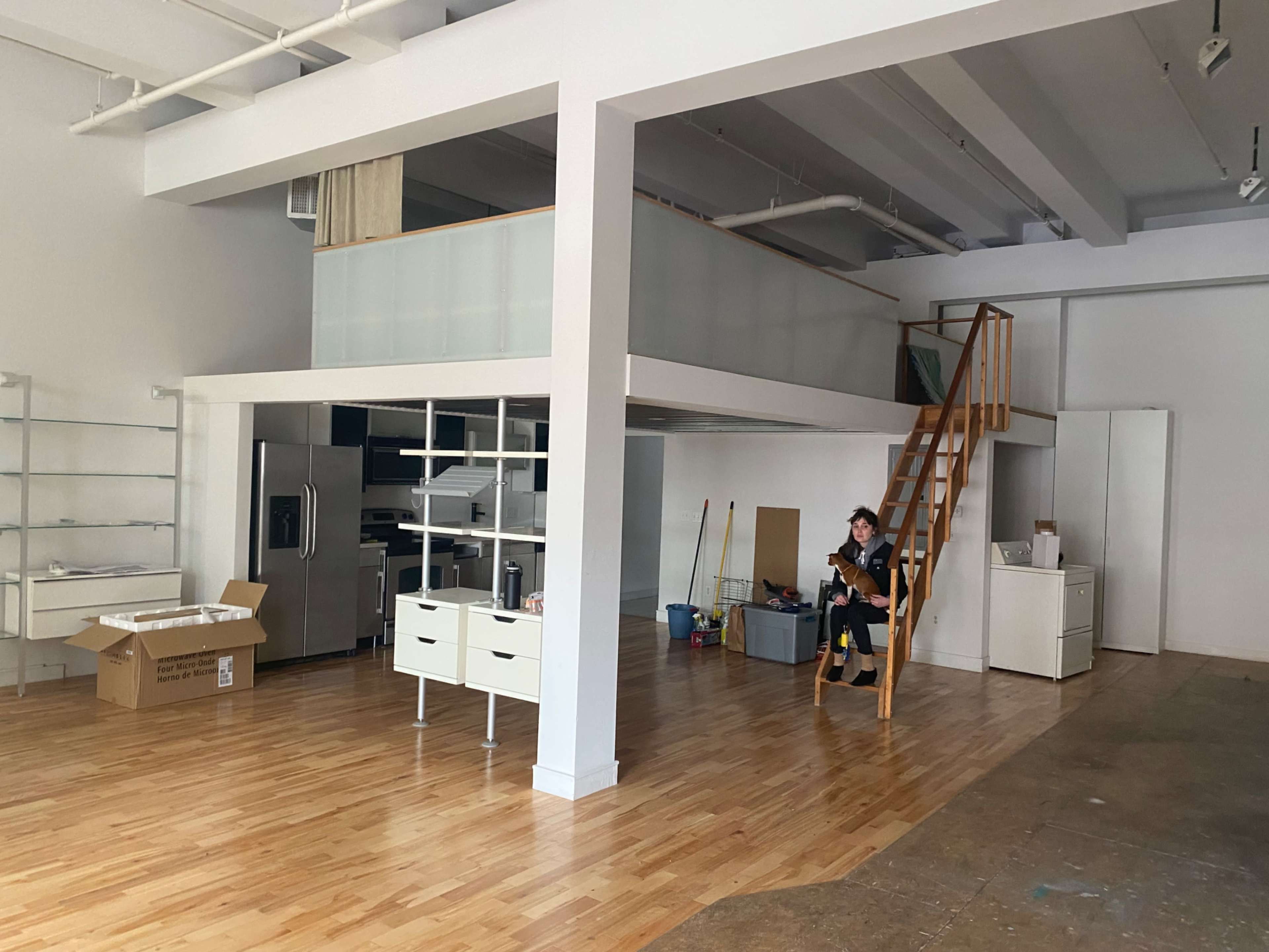 A person sits on a staircase in a spacious, partially furnished room with wooden floors and white walls.