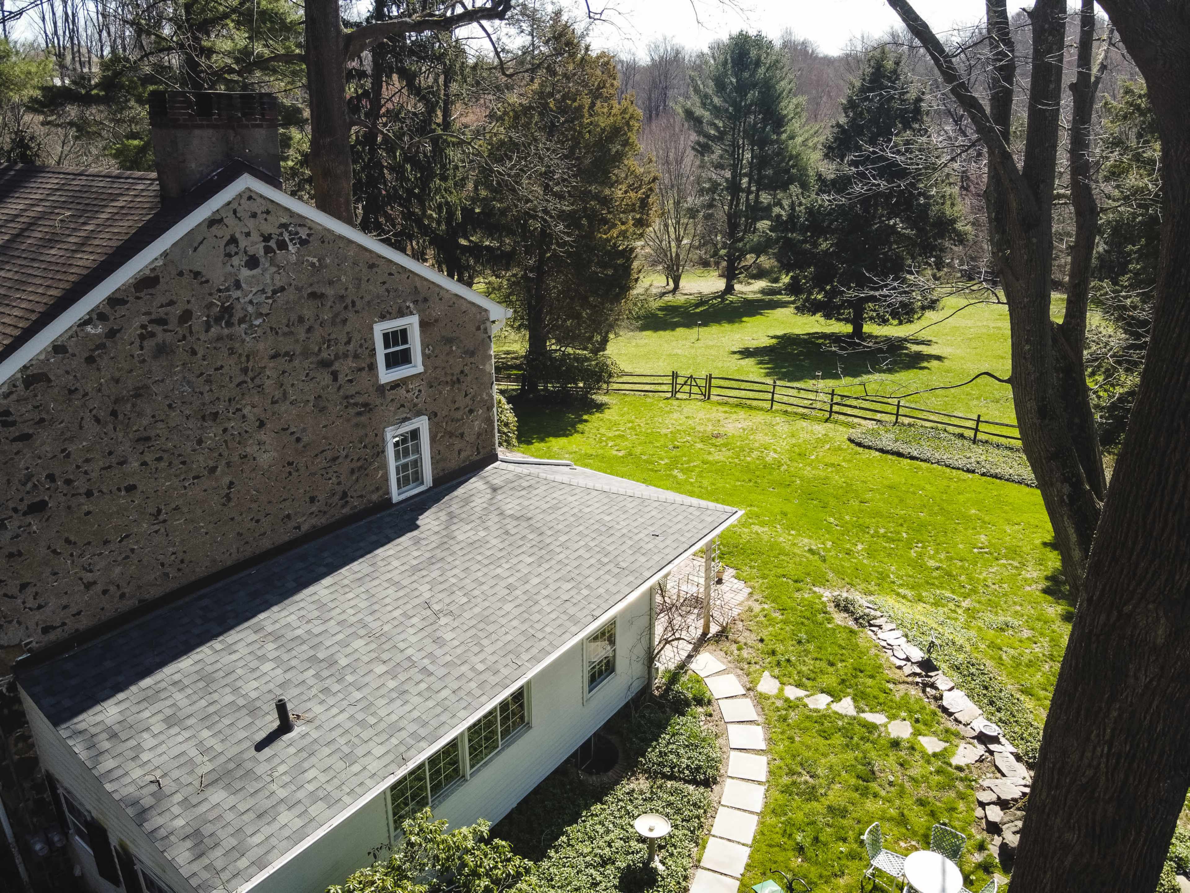 An aerial view captures a stone house with a gray roof surrounded by a green lawn, trees, and a fenced area.